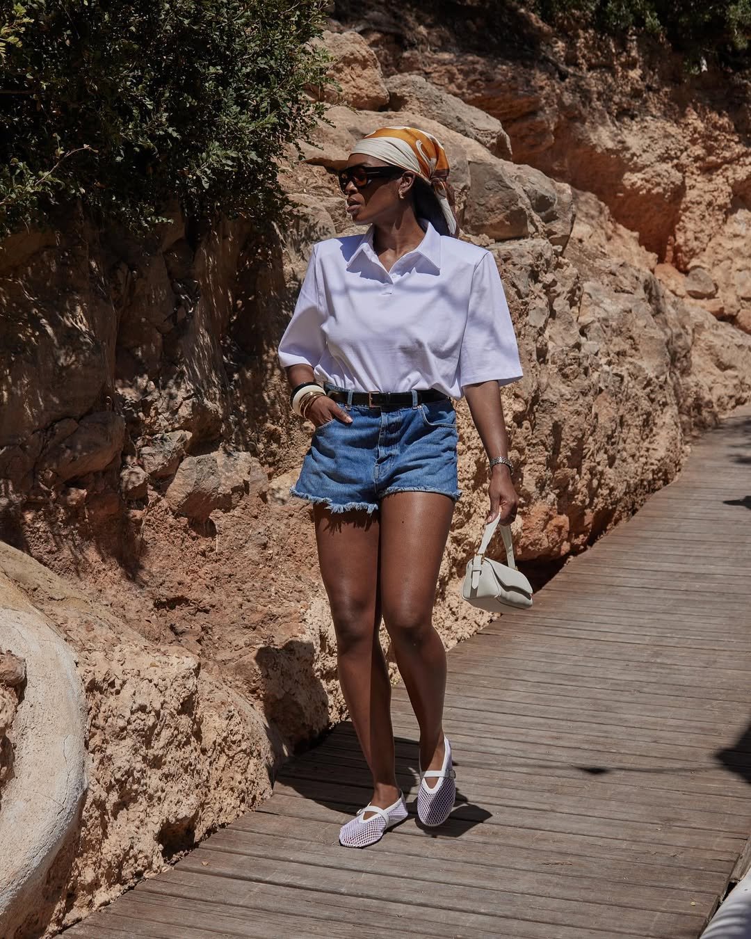 Pessoa usando blusa polo branca, shorts jeans e lenço na cabeça, caminhando em passarela de madeira ao lado de rochas.