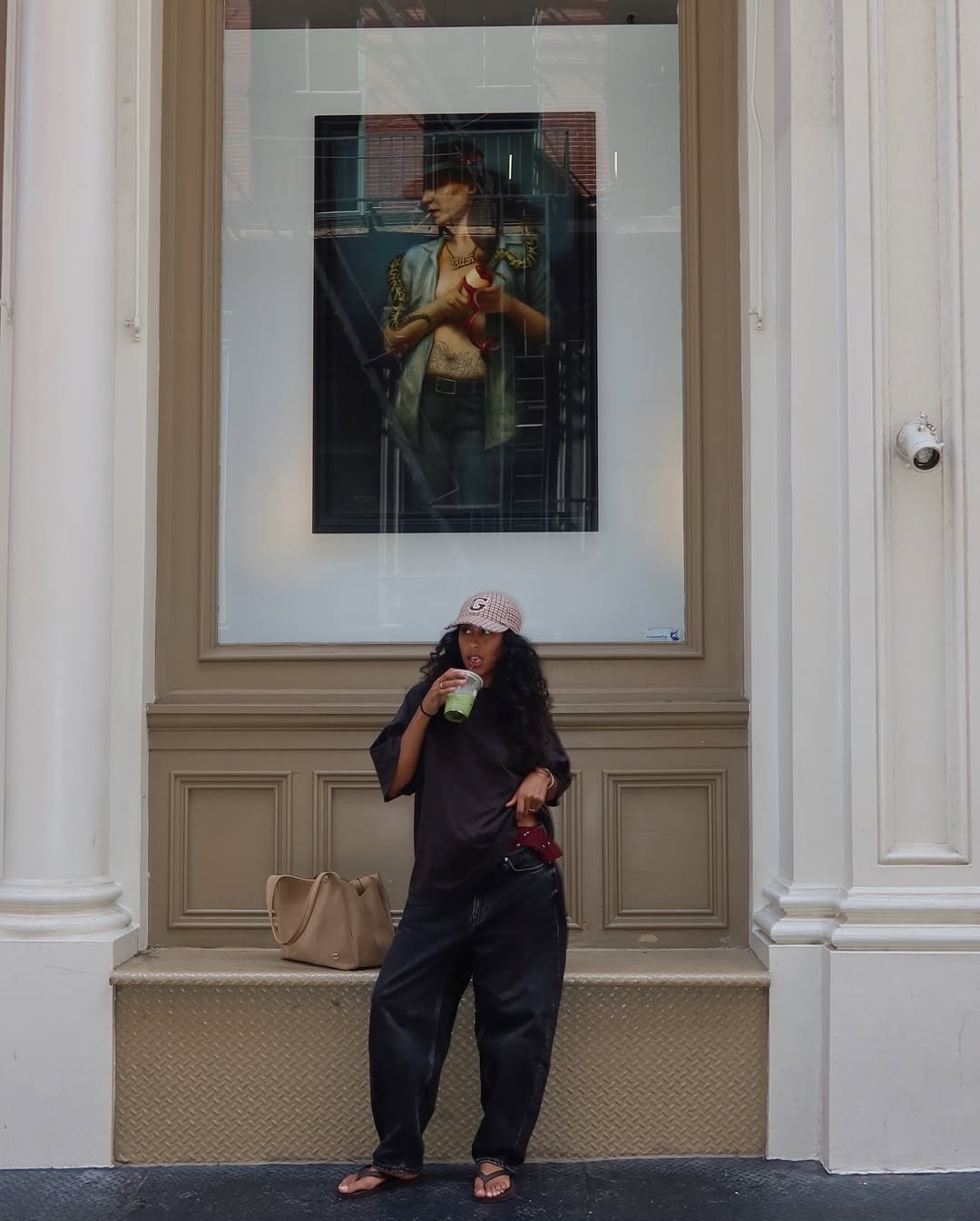 Mulher com look casual usa calça preta larga, camiseta oversized, boné estampado e chinelos, segurando bebida verde.
