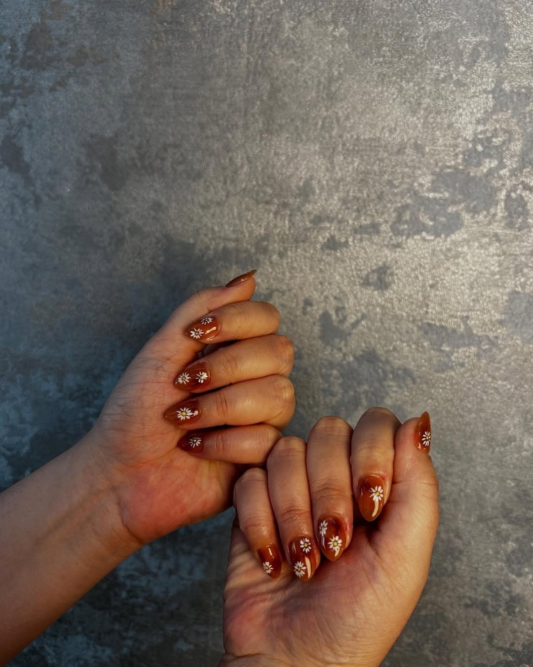 As mãos exibem unhas bem cuidadas, com esmalte em tom marrom e detalhes florais brancos.
