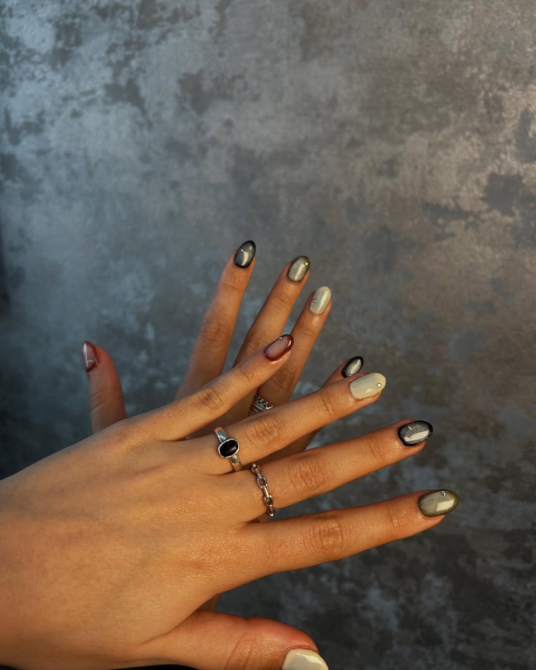 As unhas exibem uma combinação de esmaltes em tons variados, como cinza, nude e preto, com detalhes de brilho e pedras em algumas delas.