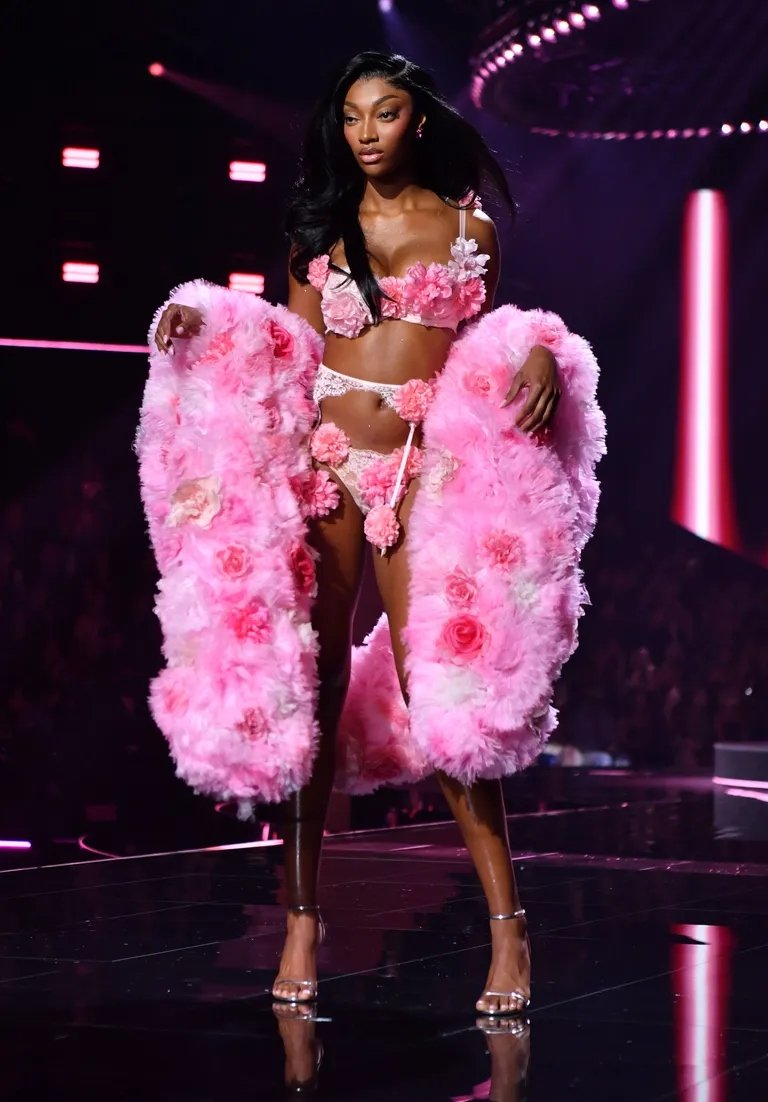 Modelo em desfile com lingerie floral rosa e casaco felpudo, exibindo penteado liso solto.