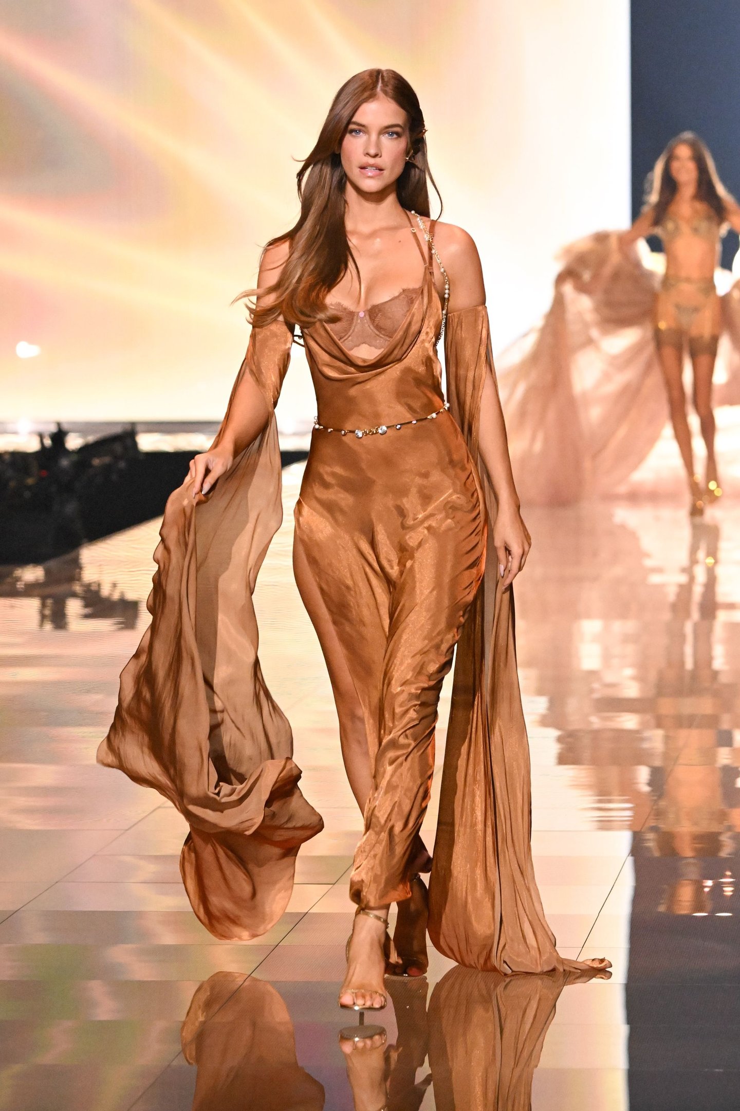 Modelo em passarela veste vestido marrom esvoaçante. Cabelos longos com penteado liso solto.
