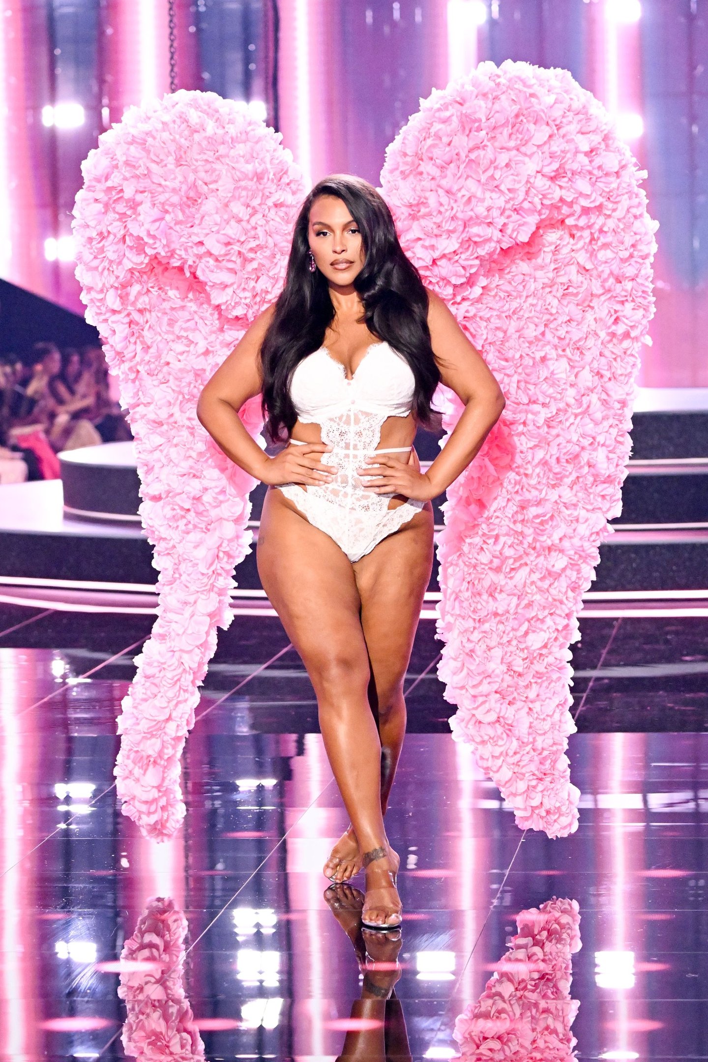 Modelo de cabelos longos em penteado liso solto e body branco, usando asas rosas em um desfile de moda.