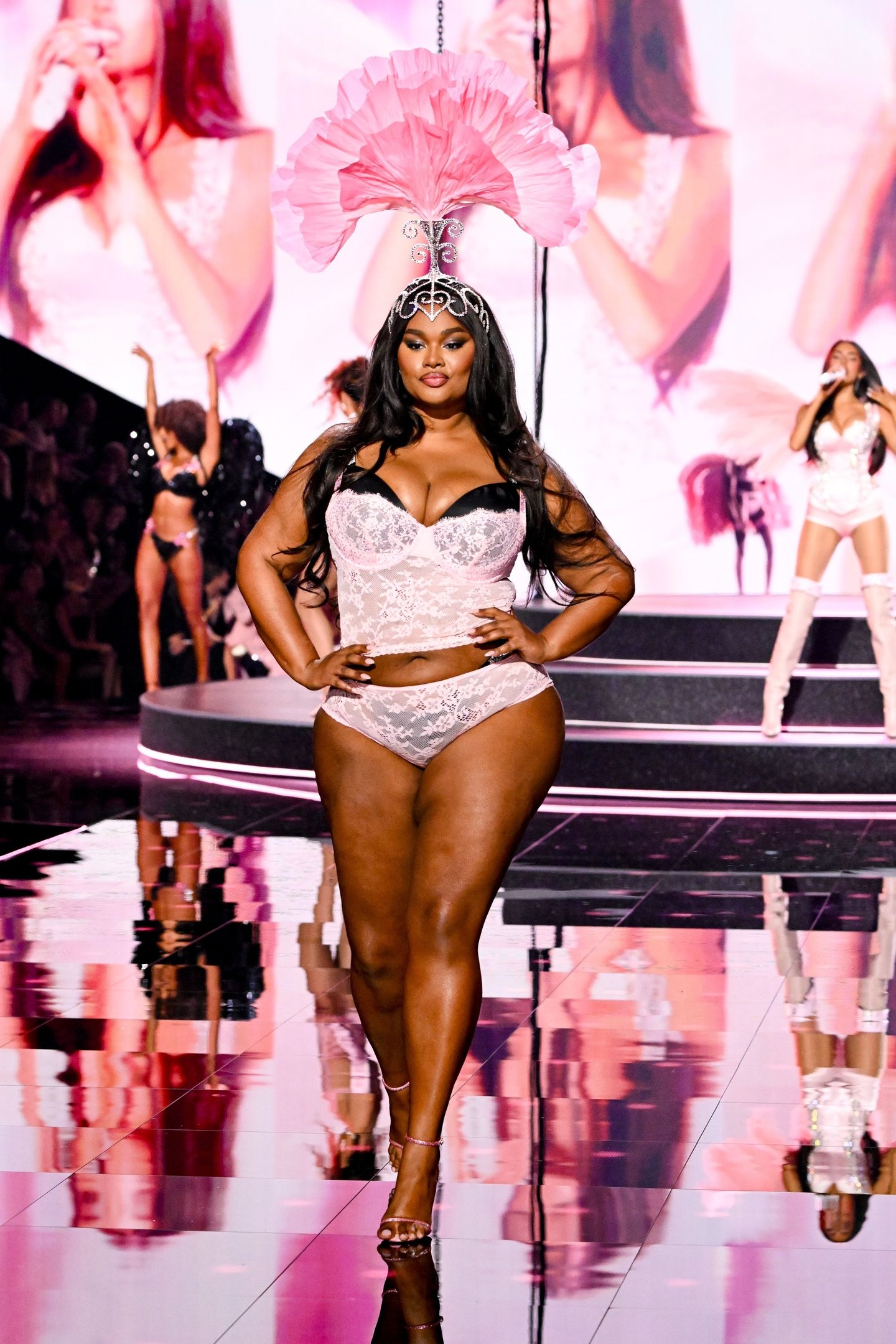Modelo com cabelo liso solto, usando lingerie rendada branca e tiara rosa com plumas na passarela.