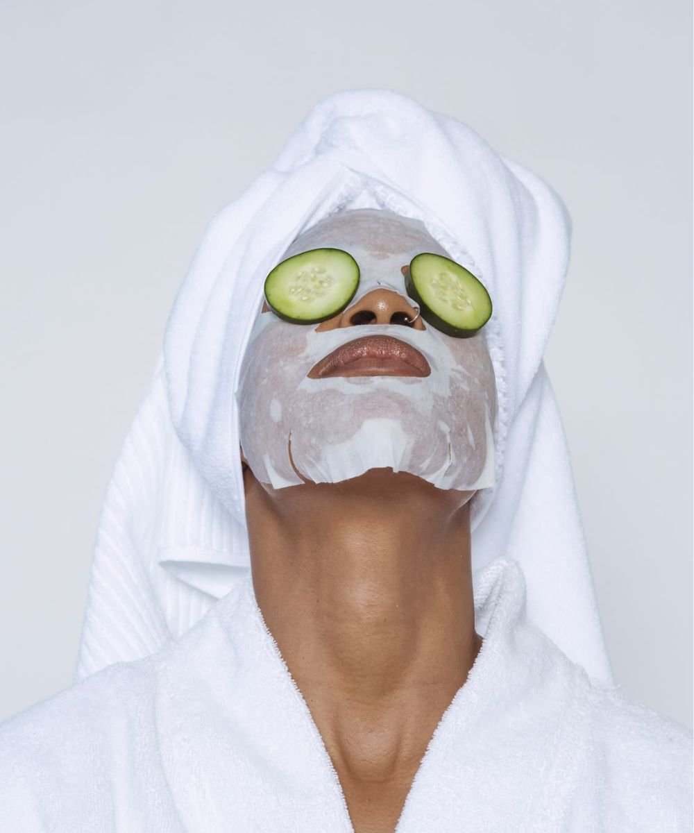 Pessoa relaxando com toalha e máscara facial, olhos cobertos por rodelas de pepino, destacando benefícios do pepino.