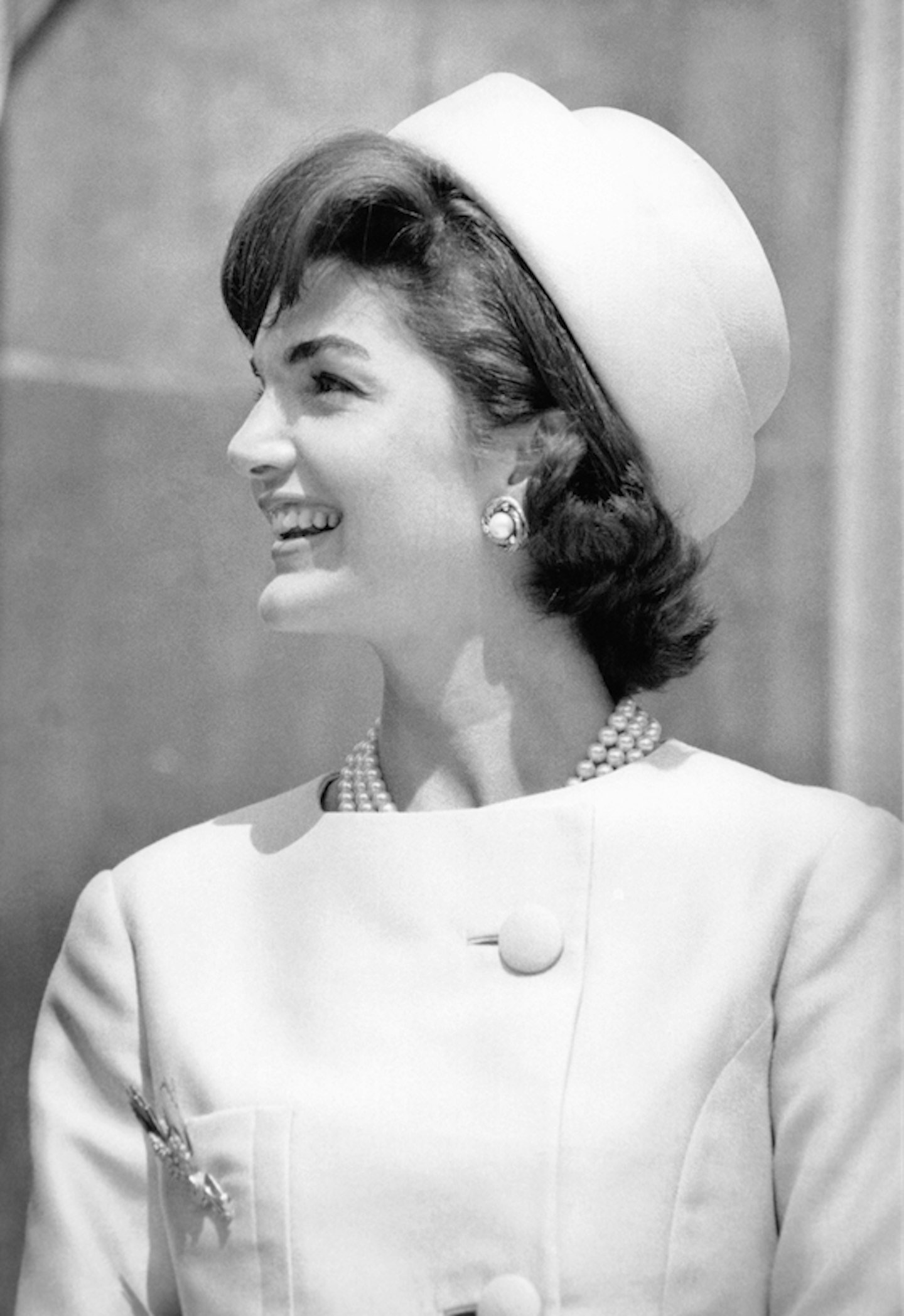 Mulher sorrindo, usando chapéu pillbox elegante, casaco de botões e colar de pérolas.