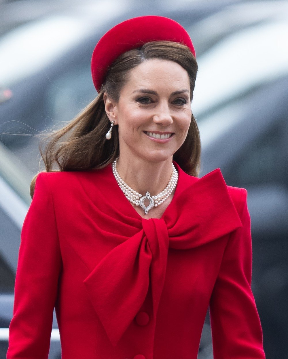 Mulher sorridente com chapéu pillbox vermelho, casaco combinando e colar de pérolas.