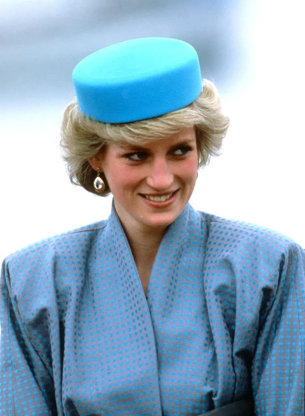 Mulher sorridente usa chapéu pillbox azul combinando com traje elegante.