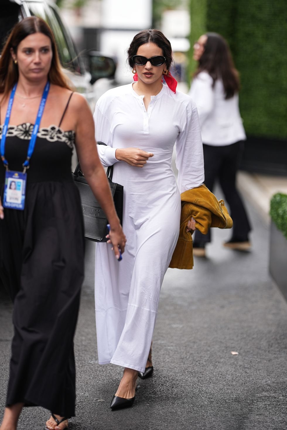 Mulher elegante de branco com óculos escuros e cabelo preso, segura casaco amarelo. Rosalía aparece em segundo plano.