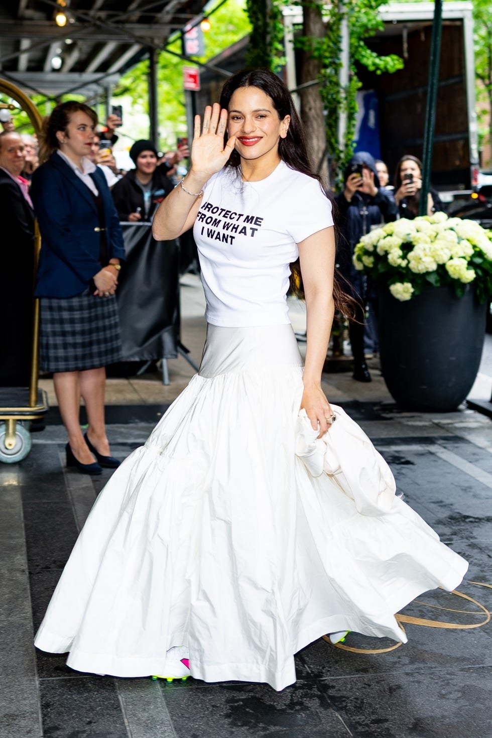 Pessoa sorridente acena usando camiseta com frase e saia longa branca. Rosalía está presente.