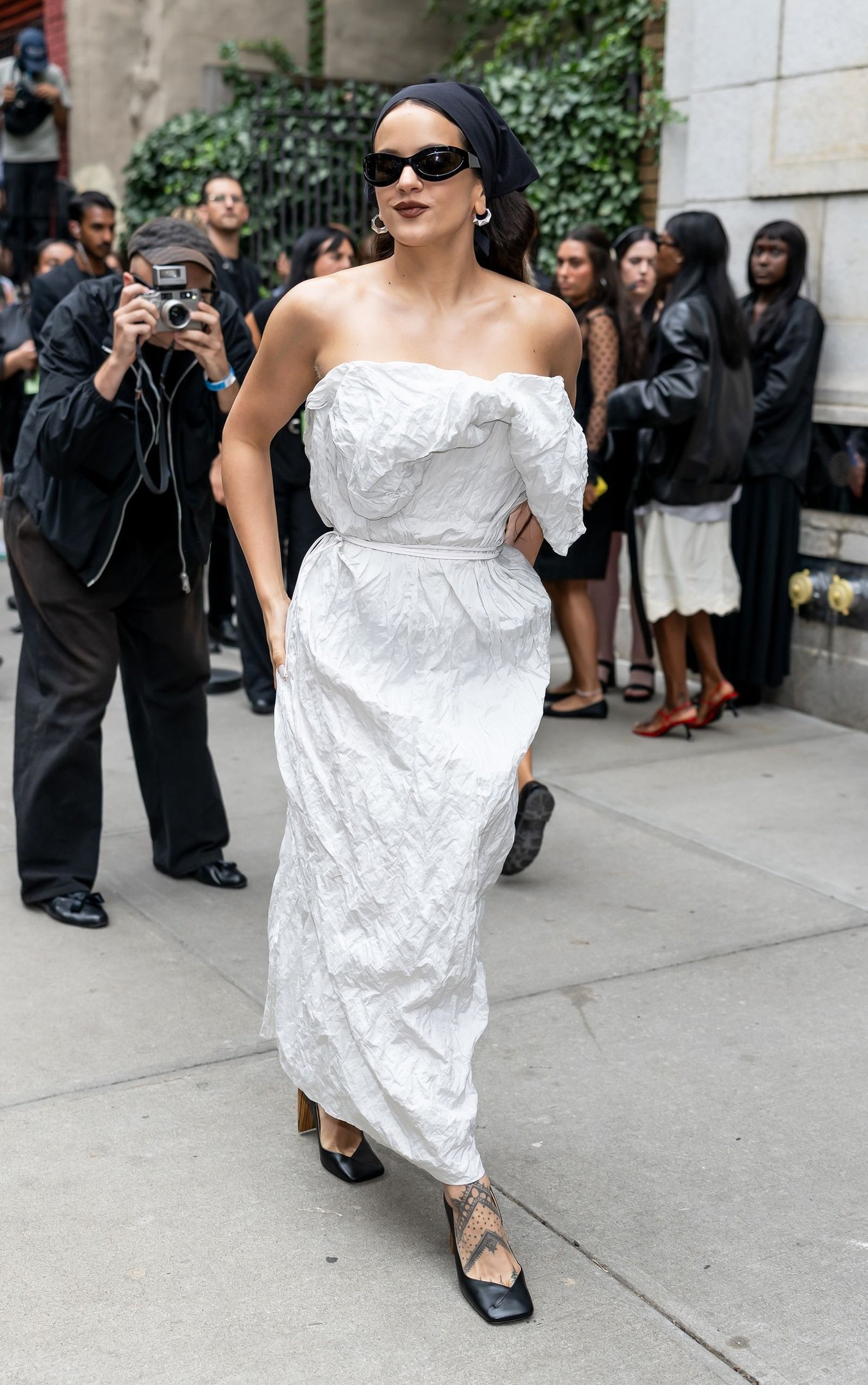 Mulher de óculos escuros e vestido branco elegante andando na calçada, cercada por pessoas.
