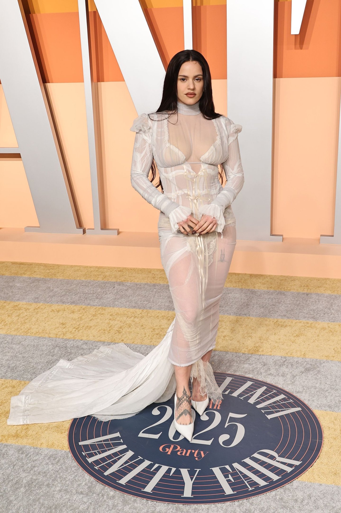 Rosalía está usando um vestido transparente branco, com cabelos longos e escuros, em um evento de tapete amarelo e cinza.