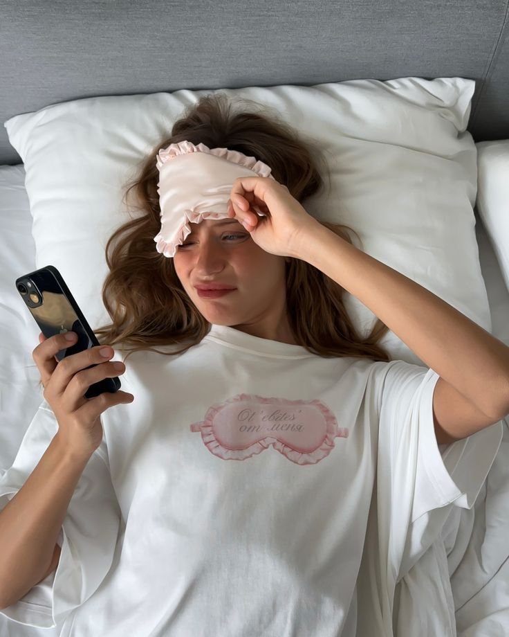 Mulher acordando na cama, olhando o celular com máscara de dormir na testa, sugerindo início do dia. Como vencer a procrastinação