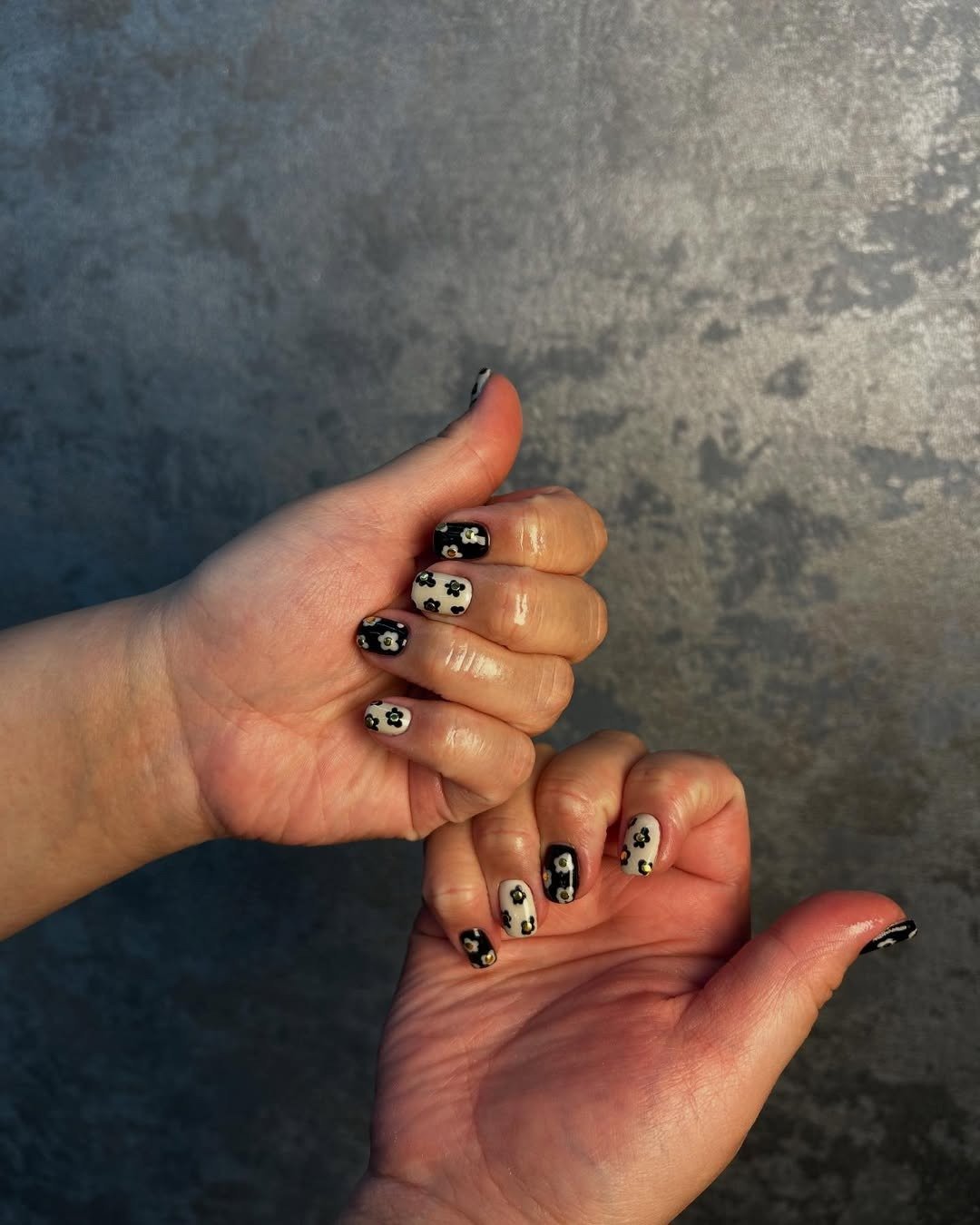 As mãos exibem unhas decoradas com esmalte descasque, apresentando um design moderno em cores preto e creme, adornadas com pequenas flores.