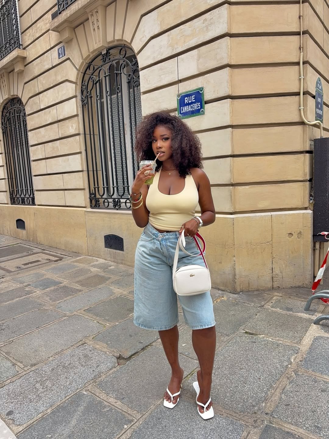 A regata curtinha, em tom nude, é a peça central deste look moderno e descontraído. Combinada com um shorts jeans de cintura alta, traz frescor e estilo. A modelo segura uma bebida, complementando o visual com um toque casual.