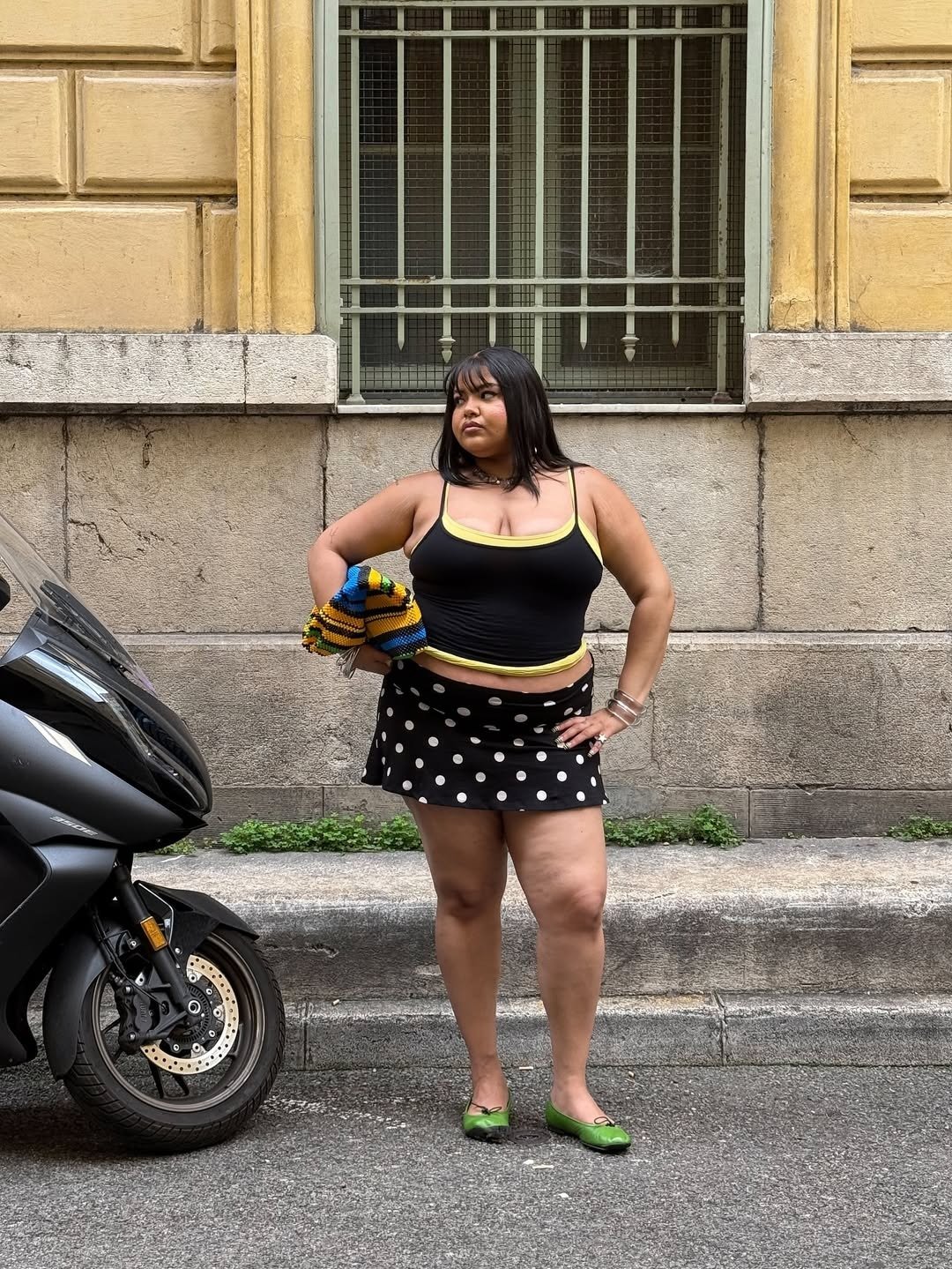 A regata curtinha preta com detalhes em amarelo complementa um look ousado, combinada com uma saia de bolinhas brancas. A modelo, com cabelo liso e acessórios marcantes, exibe confiança e estilo.