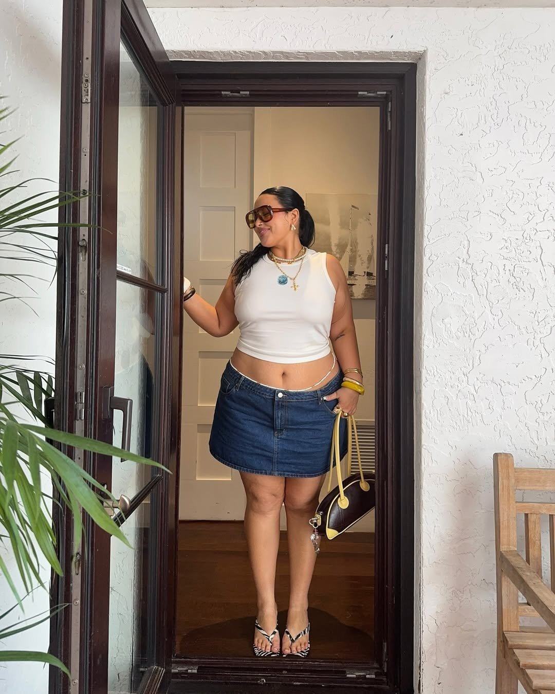 A regata curtinha branca é um destaque no look, combinando com uma saia jeans curta que valoriza a silhueta. A modelo usa acessórios como colares e pulseiras coloridas, além de óculos de sol grandes, que adicionam estilo.