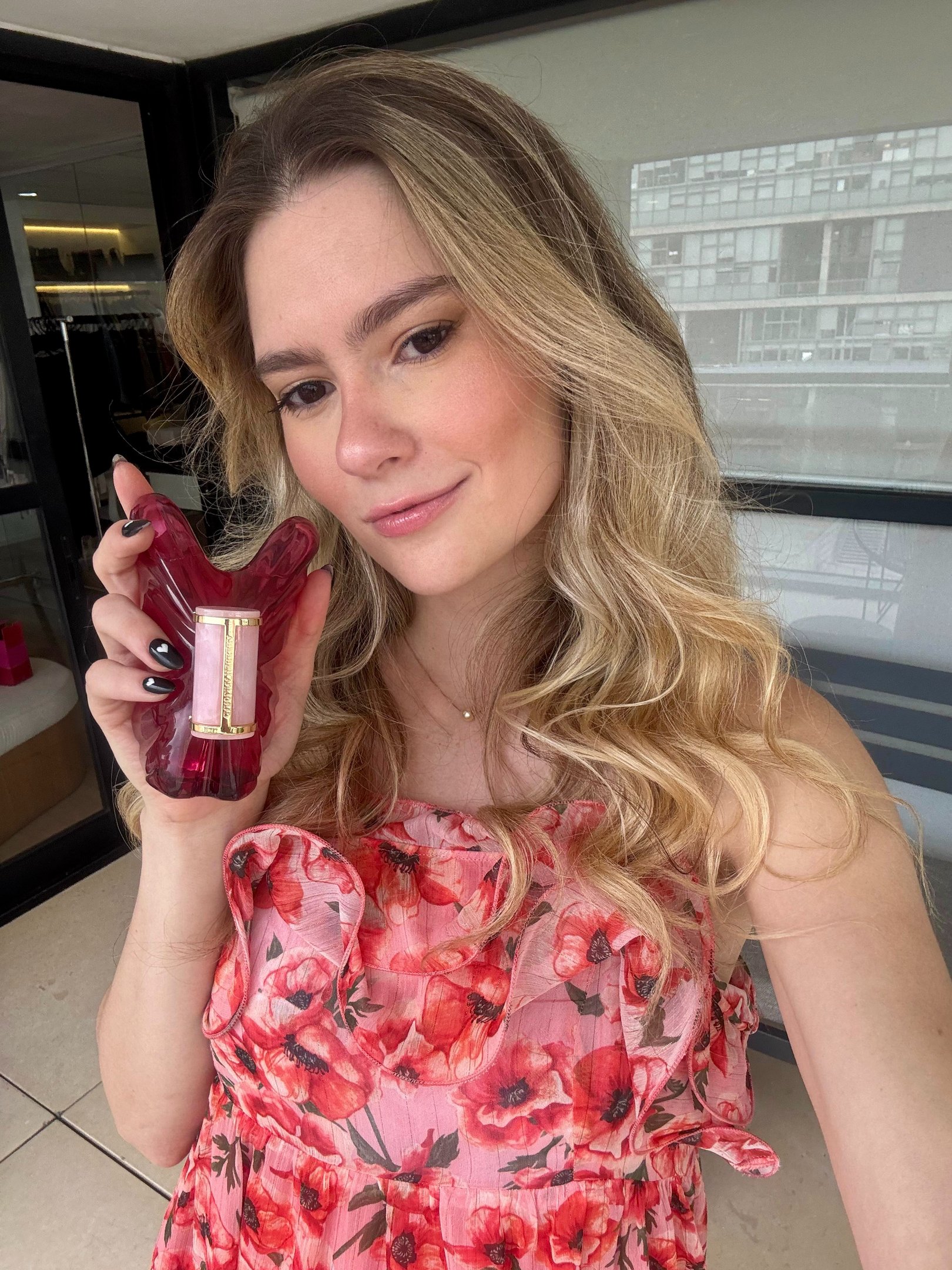 A imagem apresenta uma mulher com cabelos longos e ondulados, segurando um frasco de perfume floral para primavera. Ela veste um vestido leve com estampas de flores vermelhas, ideal para a primavera. O cenário é moderno, com janelas amplas ao fundo