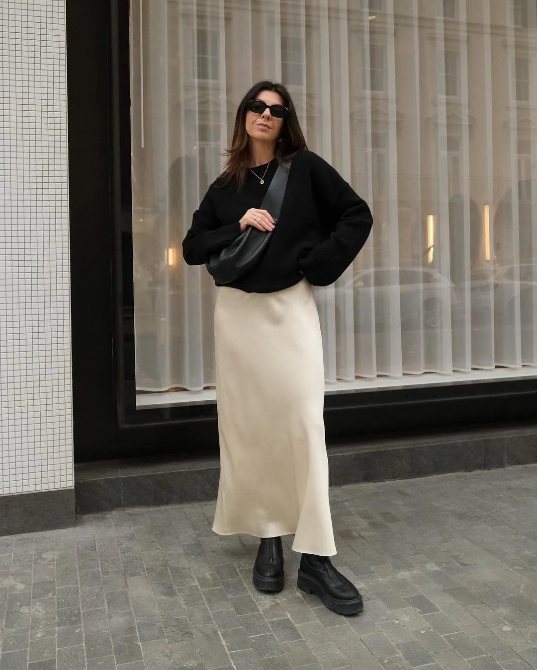 Mulher usando saia de cetim bege e suéter preto, com bolsa transversal e óculos escuros, em frente a cortina.