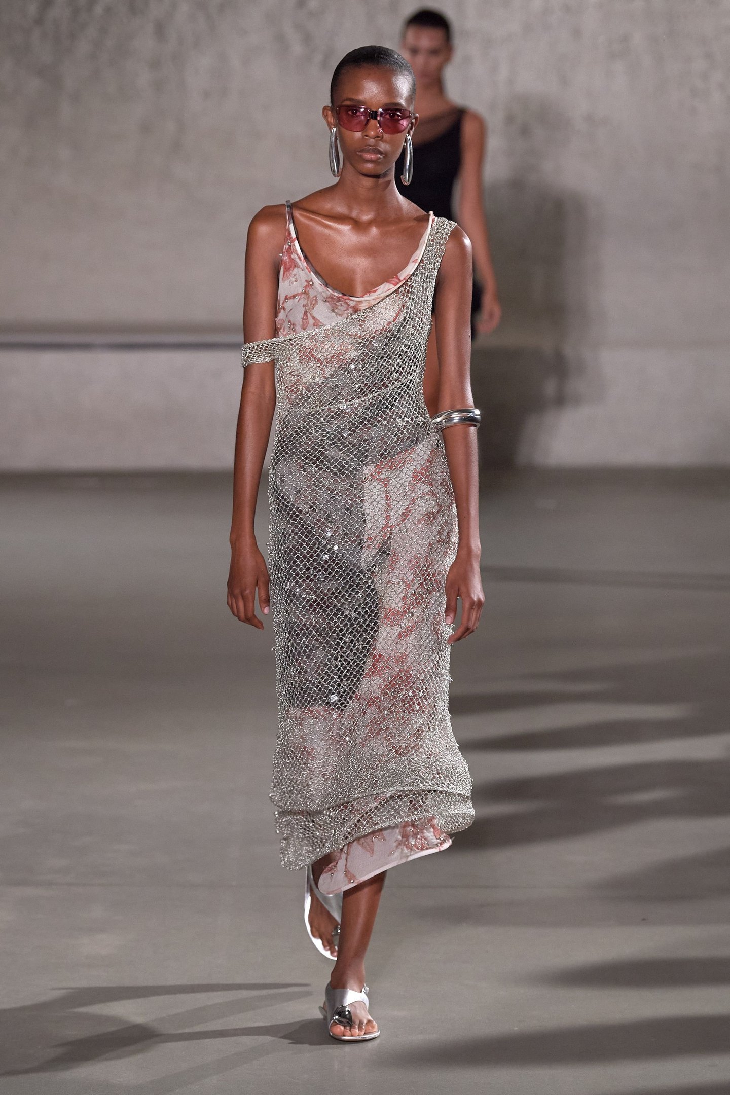 Modelo desfilando com vestido de malha prateada, par de sandálias com detalhe metálico e óculos de sol rosa.