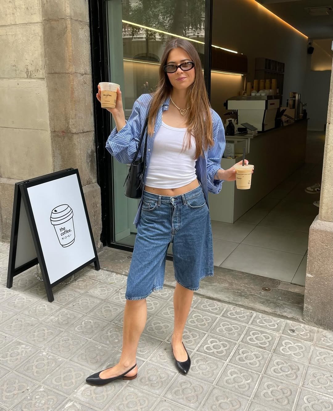 A imagem apresenta uma jovem estilosa em um ambiente urbano, segurando dois copos de café. Ela veste uma camiseta branca e uma camisa de listras azul, combinada com bermudas de jeans, criando um look casual. Modelos de sapatilhas