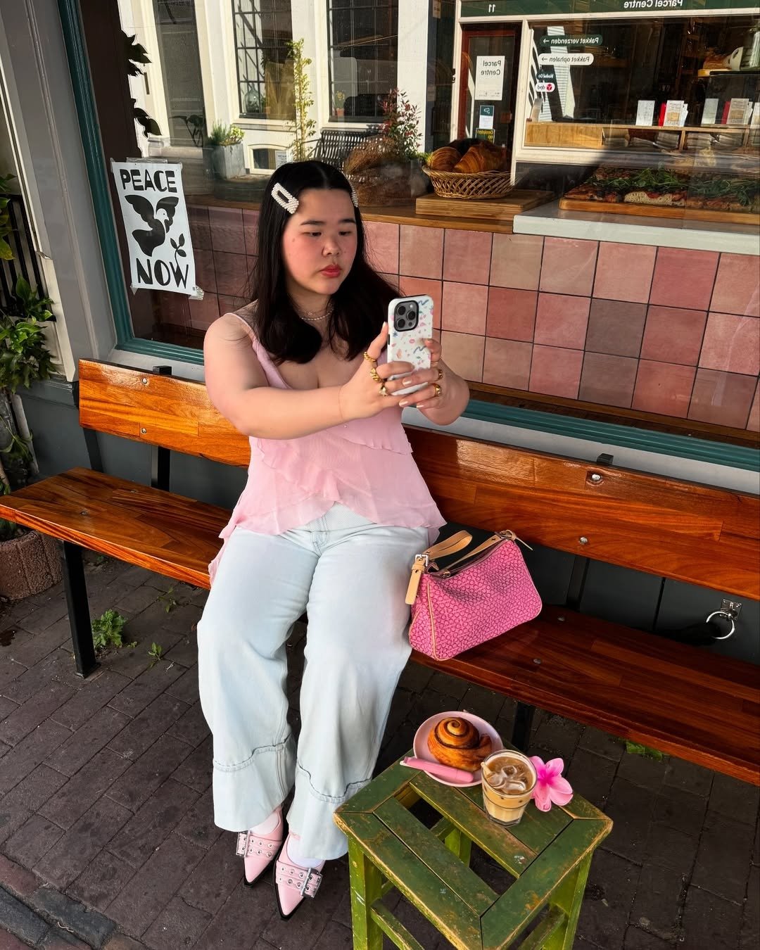 A imagem apresenta uma jovem sentada em um banco de madeira, usando uma blusa rosa de alças e calças jeans claras. Nos pés, destaca-se uma sapatilha bico fino, em tom suave, adornada com detalhes que remetem à elegância.