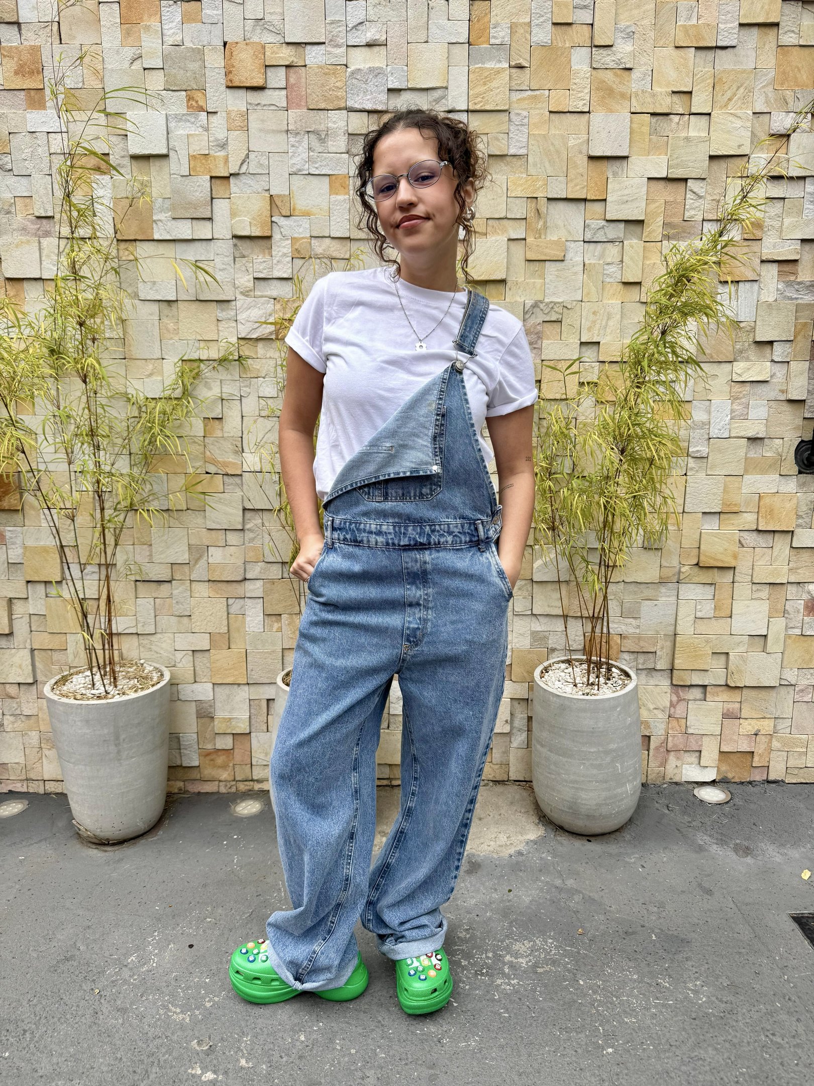 Mulher com macacão jeans e camiseta branca, destacando-se contra parede de pedra. mostrando como usar sapato colorido.
