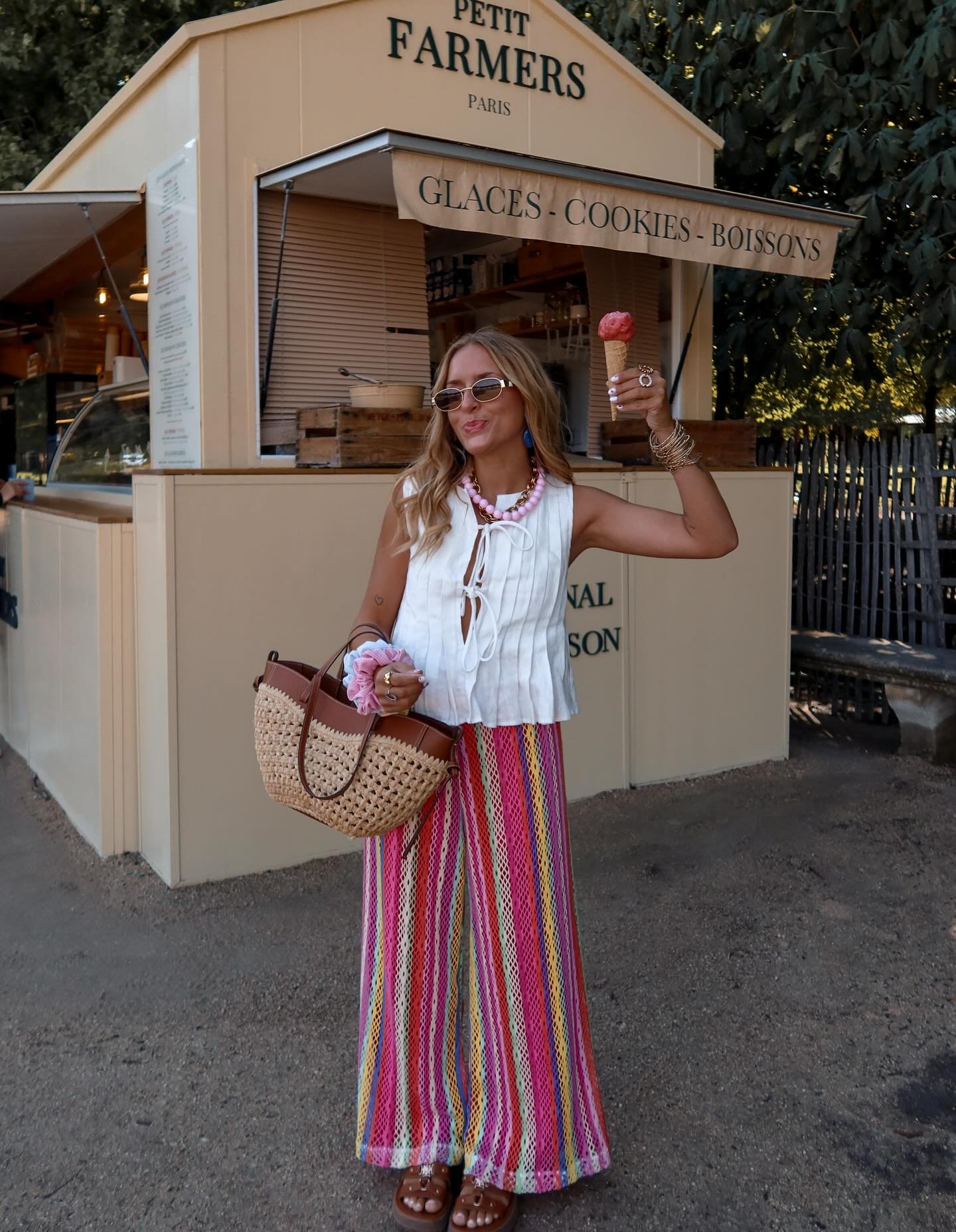 Mulher com blusa branca, calça listrada e usando sapato marrom, segurando sorvete e bolsa em frente a uma sorveteria.