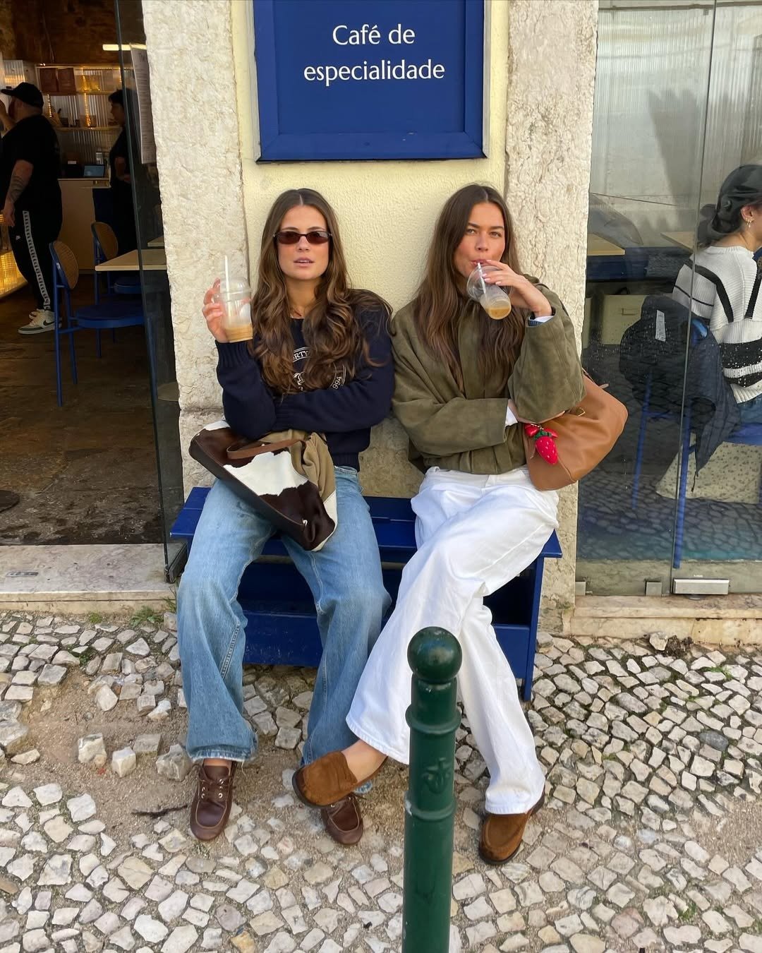 Duas mulheres estão sentadas em um banco azul, em frente a um café. Ambas usam roupas casuais e estilosas, com destaque para os sapatos tendência de 2026. A primeira usa tênis marrons, enquanto a segunda opta por um modelo em tom claro, ambos confortáveis