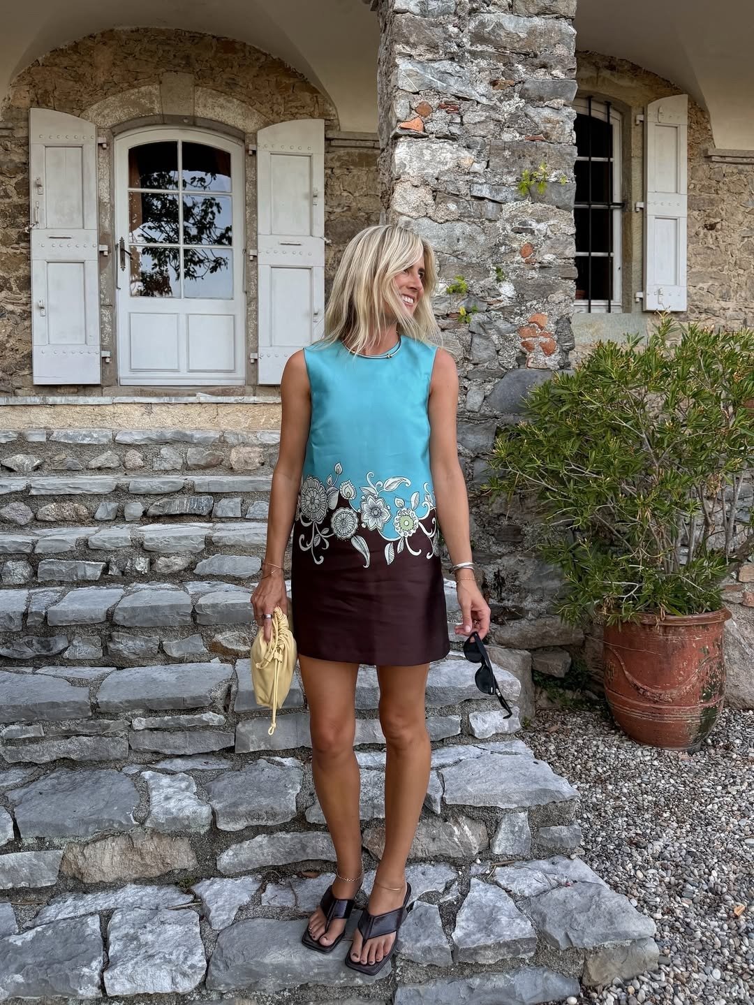 A imagem apresenta uma mulher em um ambiente rústico, com paredes de pedra e janelas brancas. Ela usa um vestido azul com detalhes florais e uma saia marrom, combinando com sapato tendência 2026.