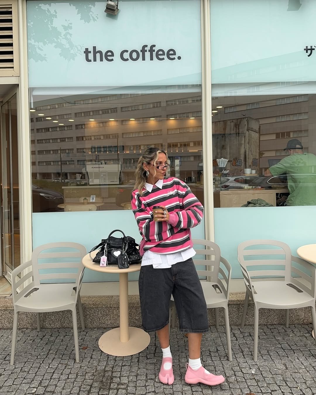 A imagem ilustra uma mulher estilosa em um café, destacando os sapatos tendência 2026, que são em tom rosa claro e com design moderno. Ela usa uma blusa listrada em rosa e preto sobre uma camisa branca, combinando com shorts escuros. A bolsa preta adicion