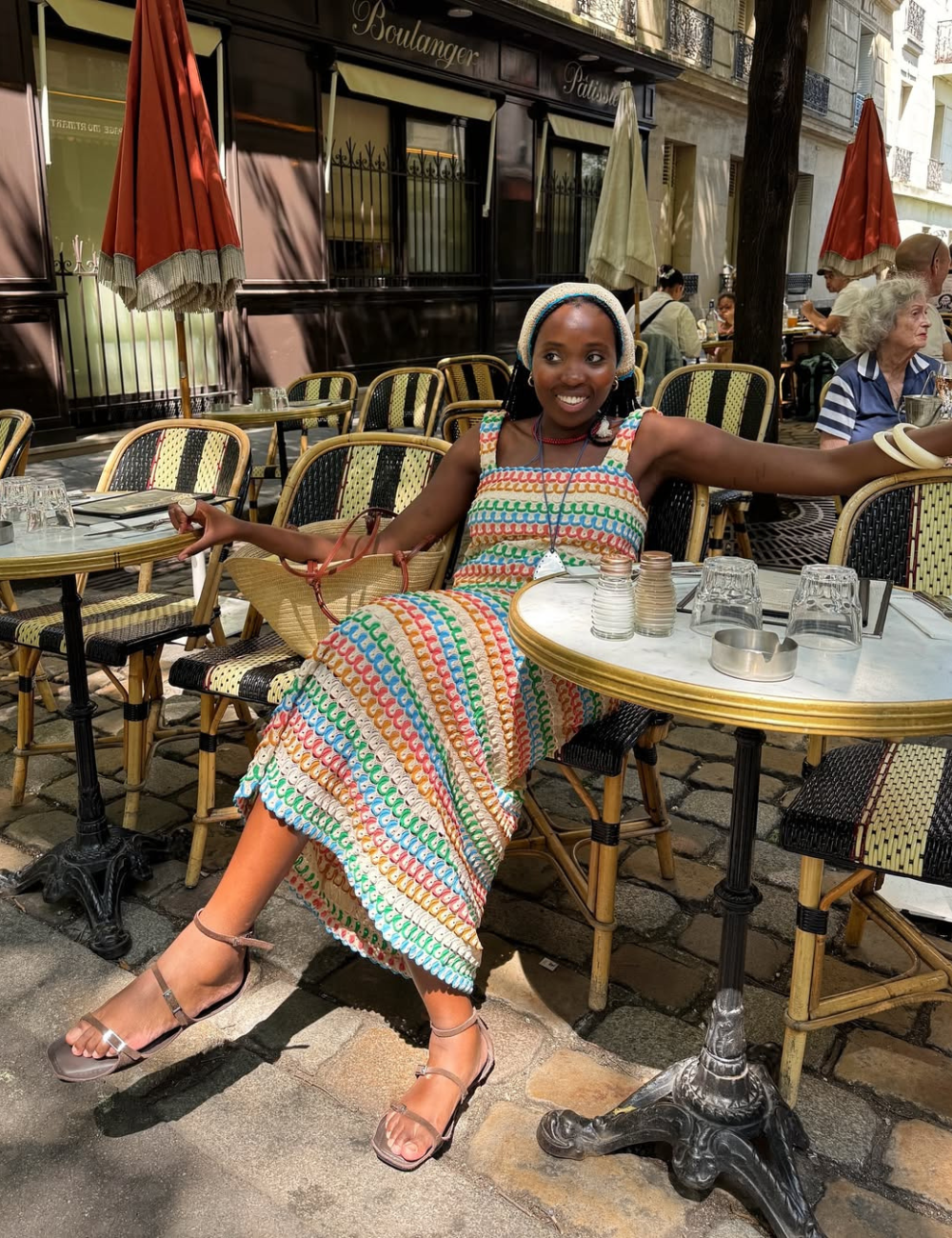A imagem destaca uma mulher sentada em um café, usando um vestido colorido de verão que remete às tendências de moda de 2026. Seus sapatos, delicadas sandálias de tiras, complementam o look fresco e elegante, ideal para climas quentes.