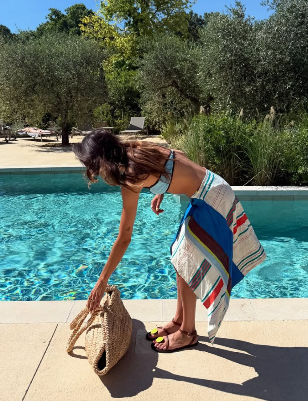 Uma mulher está próxima a uma piscina, usando um biquíni e uma canga com listras coloridas. Ela se inclina para pegar uma bolsa de palha. Nos pés, destaca-se um par de sapatos verão 2026, com design moderno e detalhes em cores vibrantes.