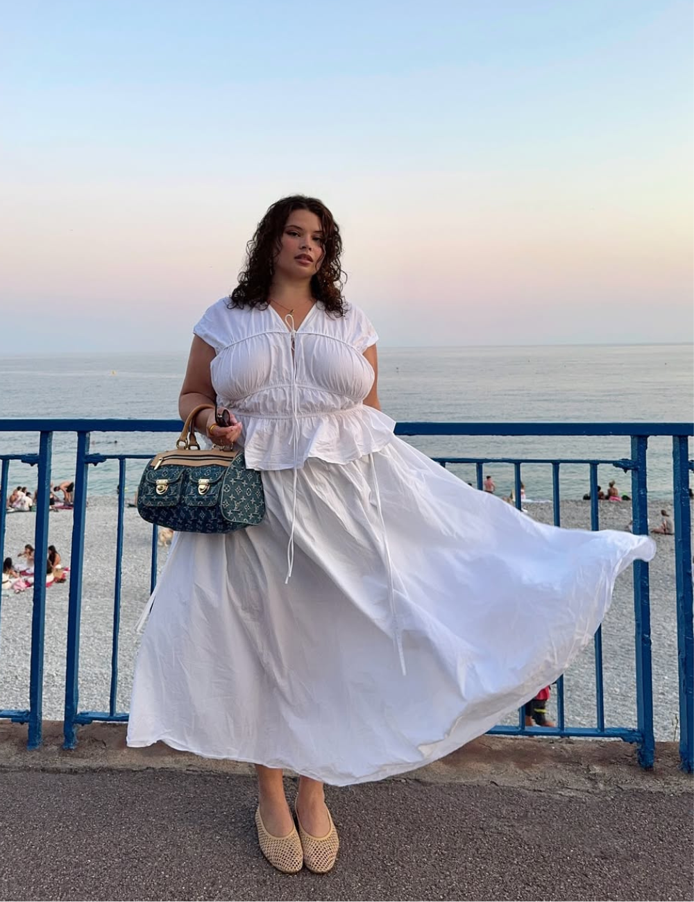 A imagem apresenta uma mulher em um cenário de praia ao entardecer, vestindo um vestido branco fluido que evoca leveza e frescor, ideal para os sapatos do verão 2026. Ela usa sapatos de palha, que combinam conforto e estilo. A bolsa de designer acrescenta