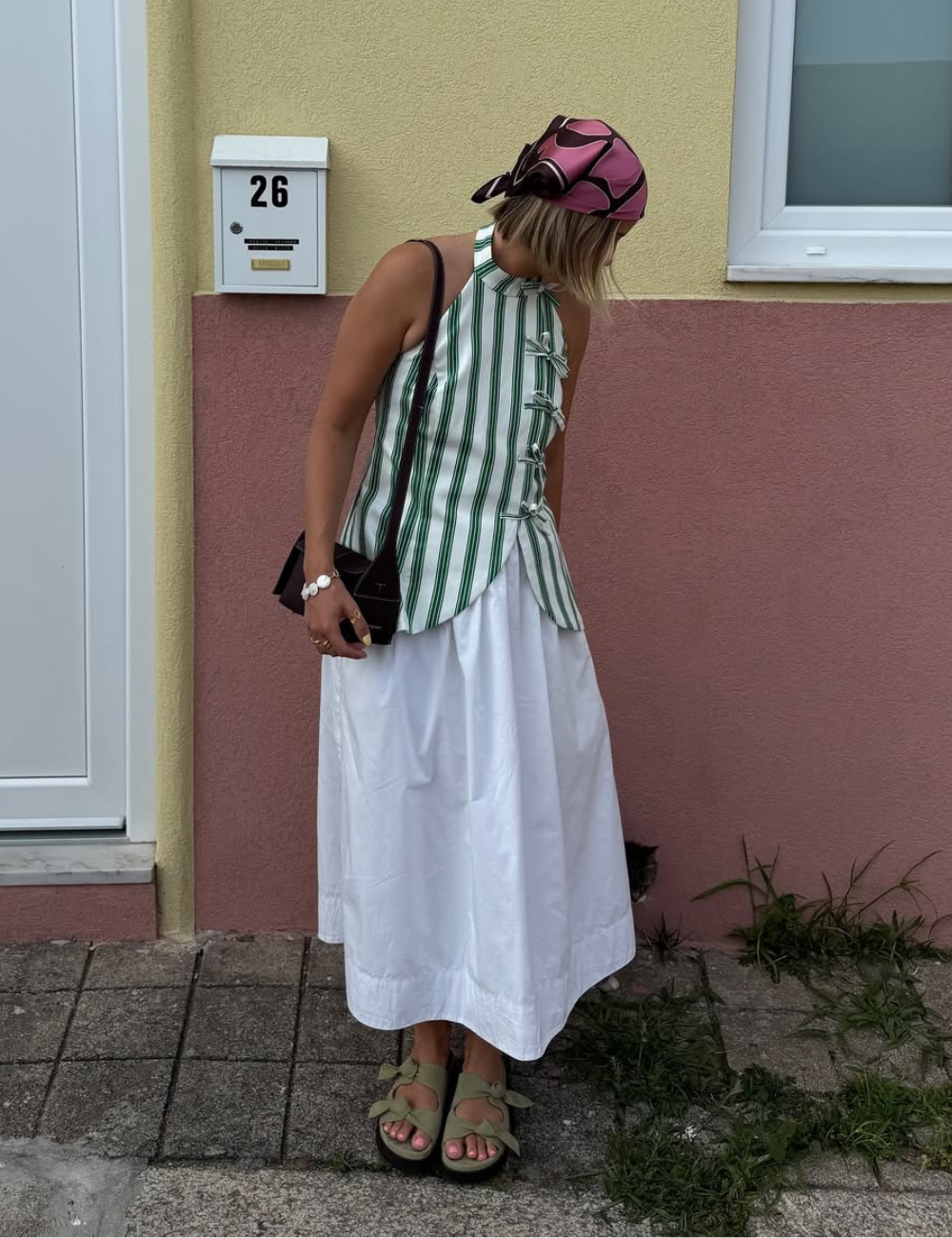 O look apresenta uma combinação de moda leve e descontraída para o verão 2026. A blusa listrada em verde e branco, com detalhes em laço, complementa a saia longa branca, criando um visual fresco.