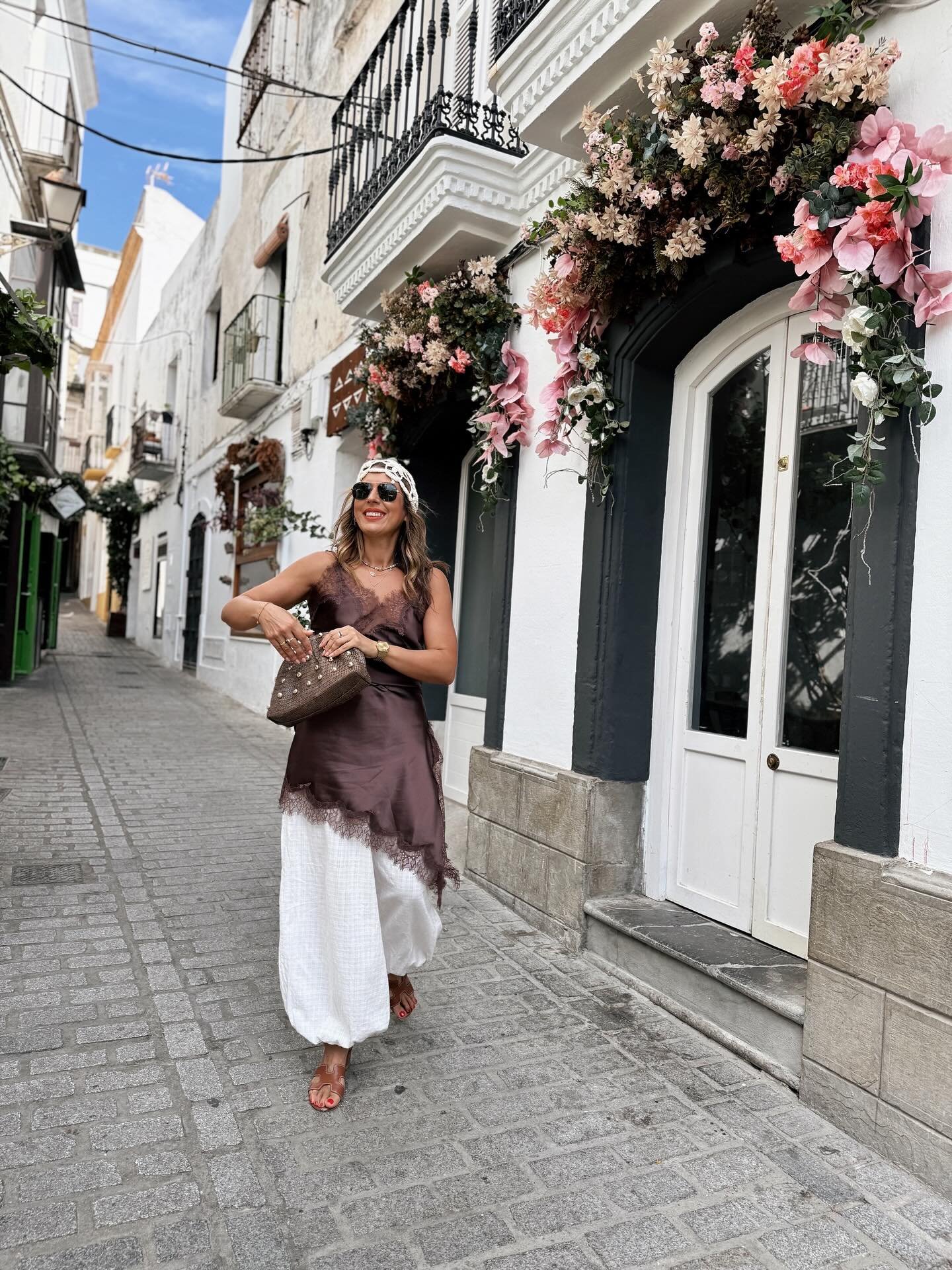 Mulher caminha em rua com vestido marrom e calças brancas, segurando bolsa marrom, perto de fachada florida usando sapato marrom