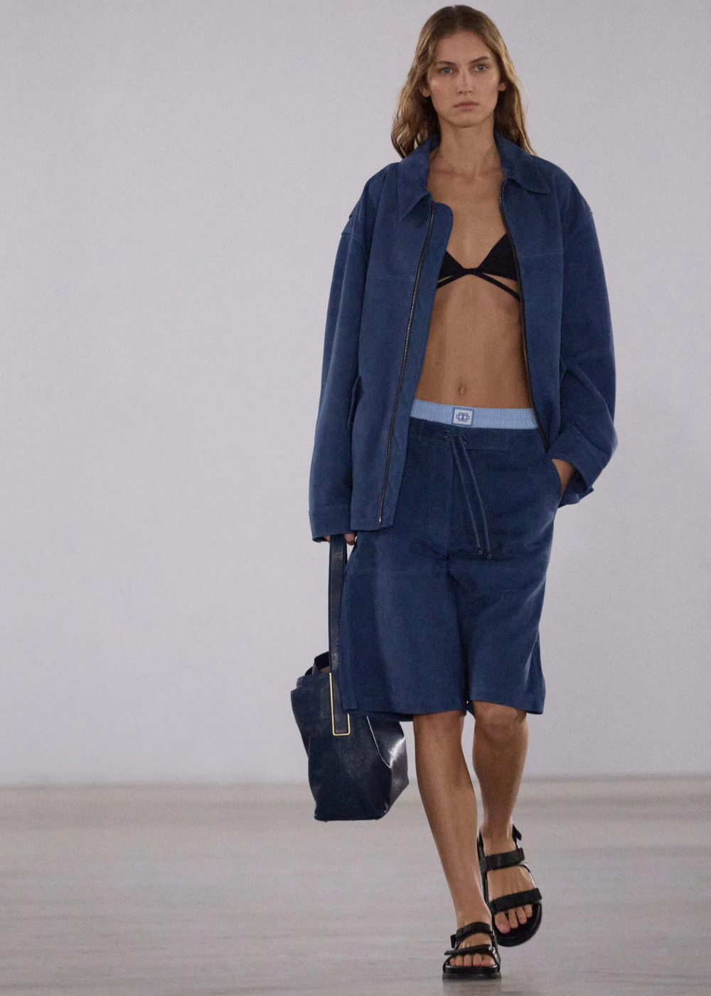 Modelo em passarela vestindo jaqueta e bermuda azul com top preto. O look remete ao estilo surf na moda.