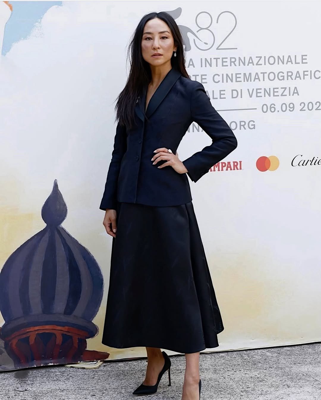 Greta Lee exibe um look sofisticado e moderno, com um blazer preto de corte estruturado e uma saia longa que complementa a silhueta. O conjunto é finalizado com sapatos de salto alto, trazendo elegância ao visual. Seu cabelo liso e solto e a make minimali