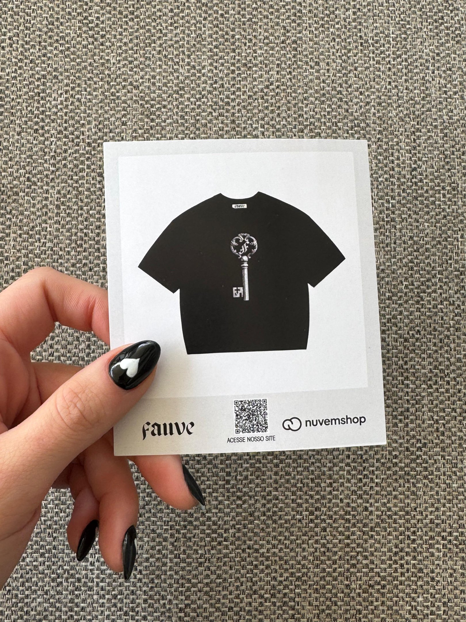 A imagem apresenta um cartão com uma ilustração de uma camiseta preta, destacando um design moderno. A peça possui uma estampa de uma chave estilizada, representando a coleção da SPFW N60. A mão que segura o cartão tem unhas com esmalte preto e um detalhe