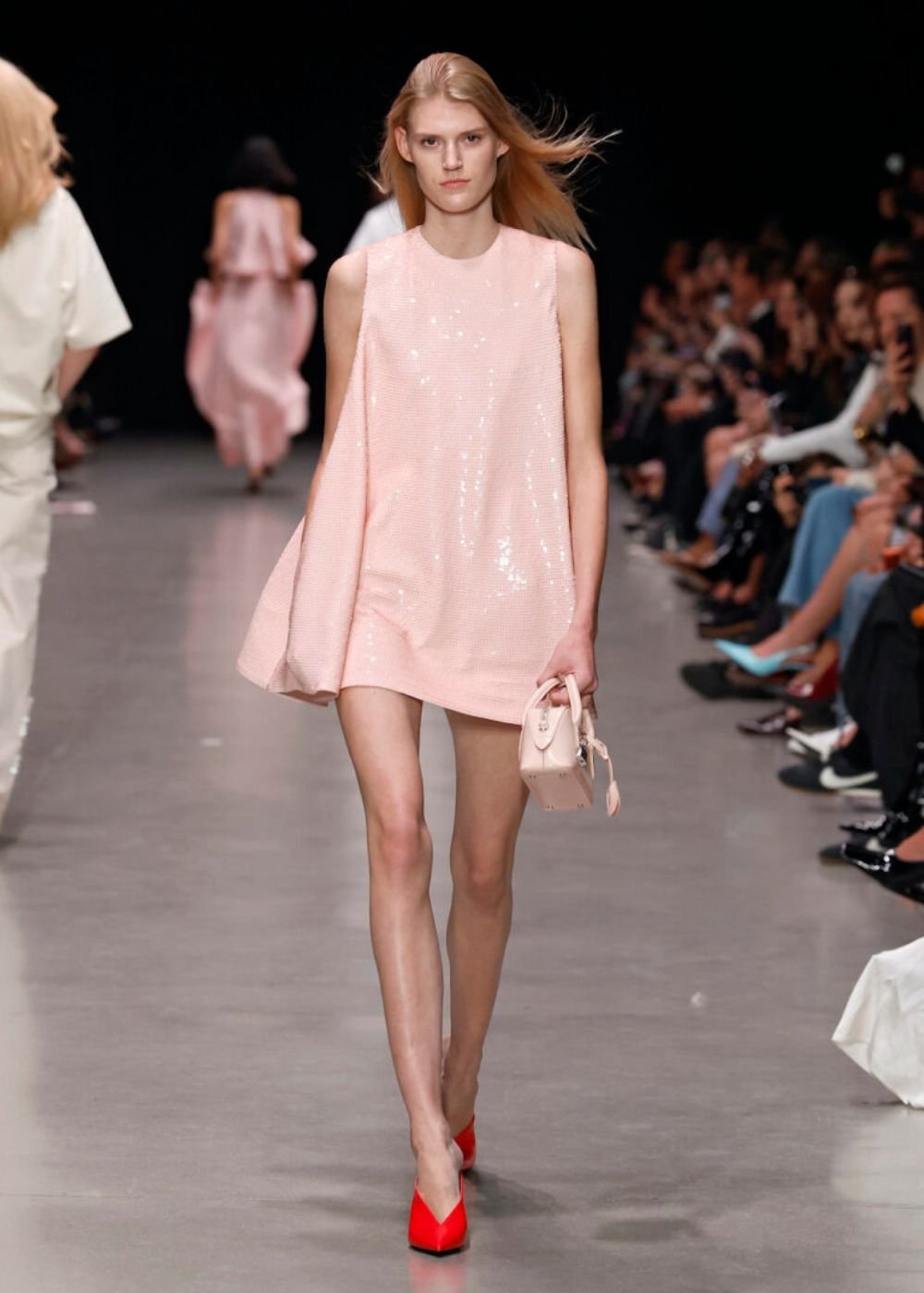 Modelo na passarela com vestido rosa bebê e bolsa combinando, usando sapatos vermelhos. Público ao fundo.
