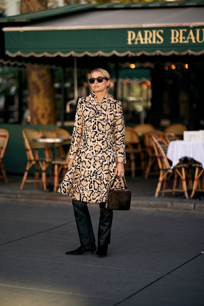 Pessoa com casaco de estampa animal, botas pretas e bolsa, exibindo street style da PFW em frente a um café parisiense.