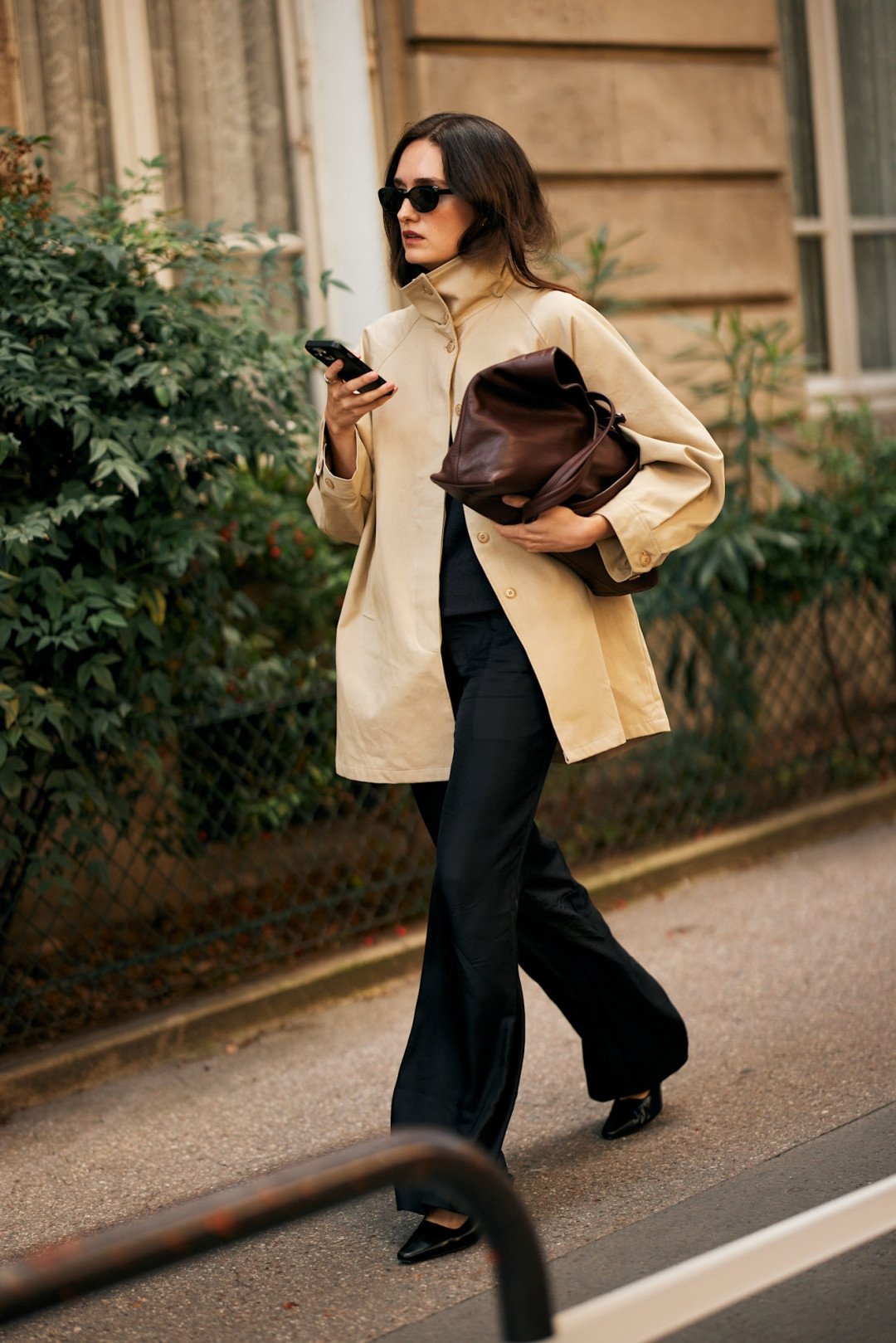 Mulher com casaco bege e óculos escuros segura bolsa marrom em look street style da PFW.