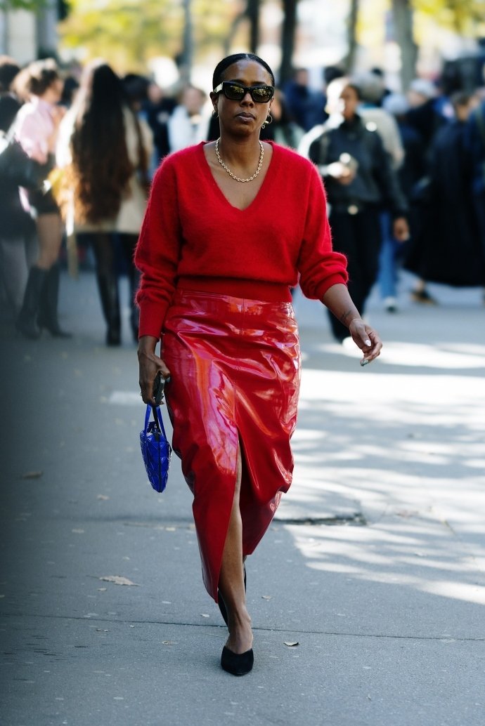 Mulher em look vermelho vibrante e bolsa azul mostra street style da PFW, com óculos escuros e colar dourado.