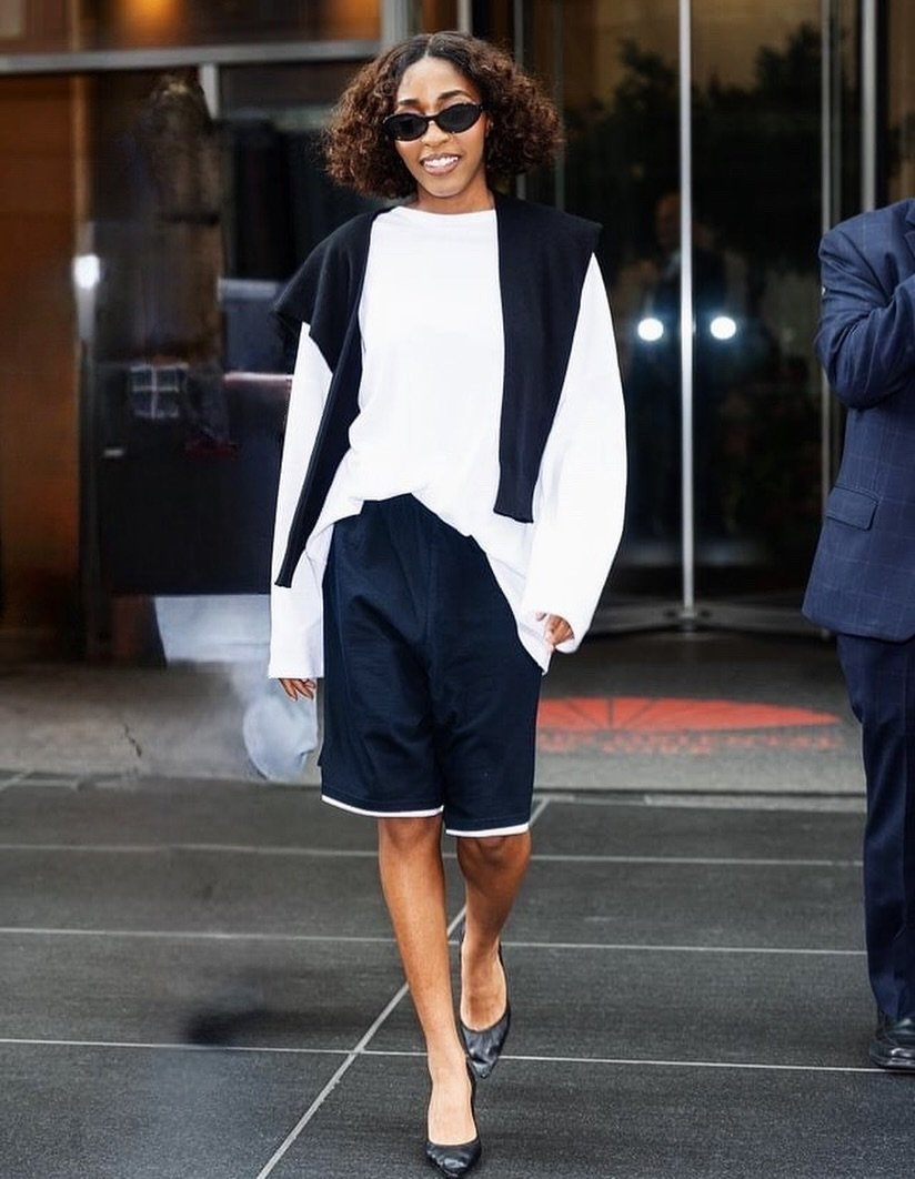 Pessoa sorrindo, usando óculos escuros, blusa branca e shorts estilo surf, caminhando em ambiente urbano.