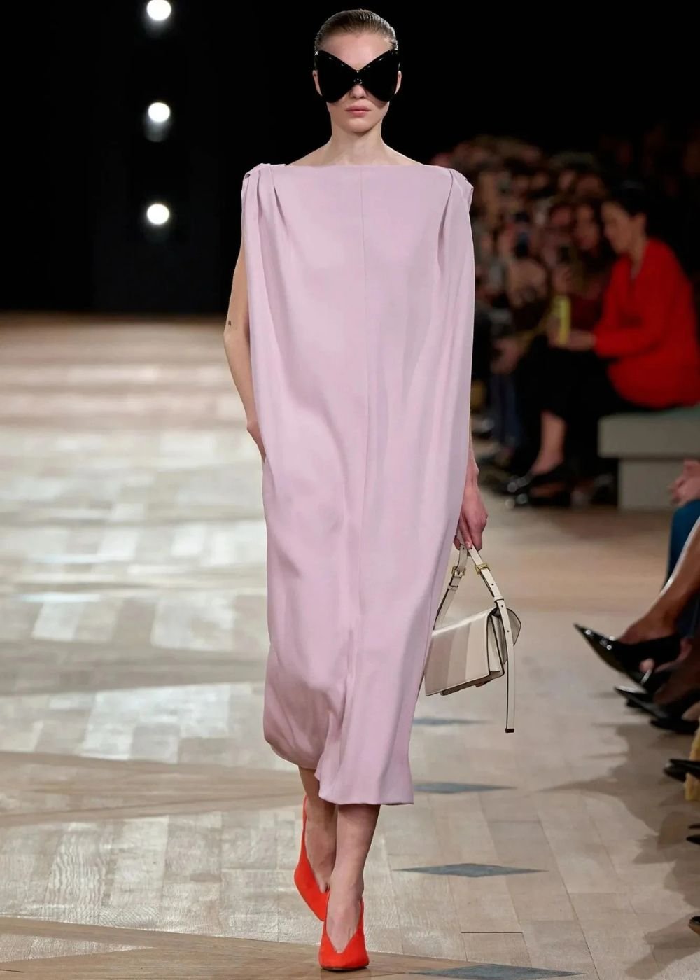 Desfile de moda com vestido reto em tom rosa suave, óculos grandes e bolsa branca.
