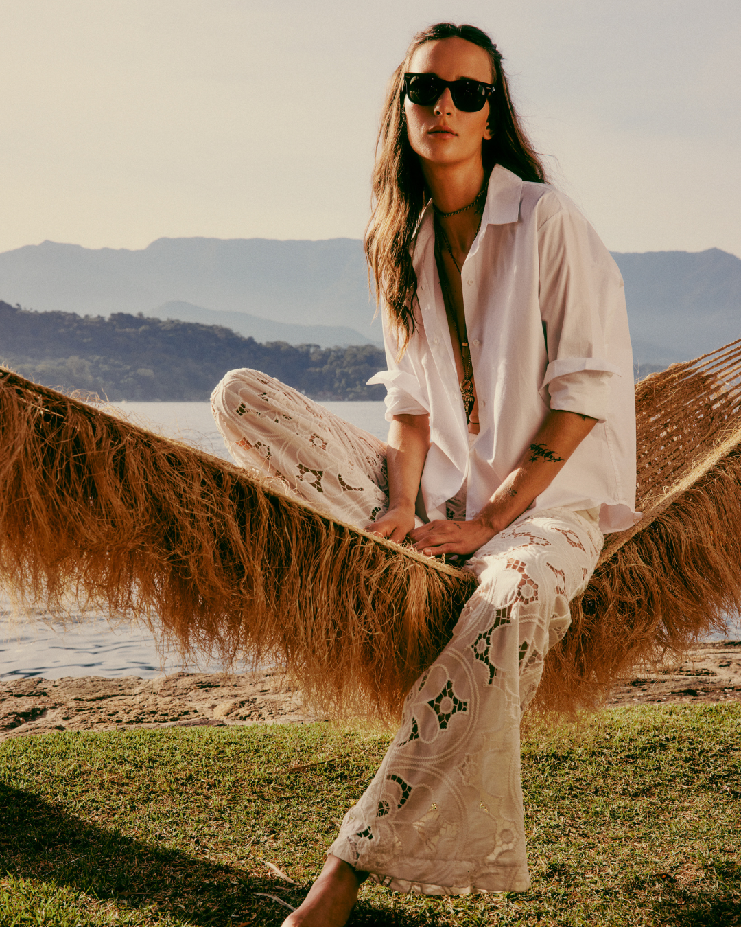 Pessoa em rede de balanço usando camisa branca e calça rendada. Look inspirado em tendências de alto verão tropical. Água e montanhas ao fundo.