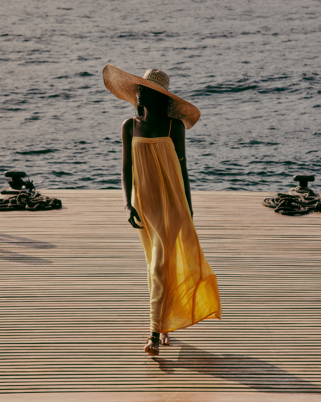 Vestido amarelo fluido e chapéu de palha usados à beira-mar, refletindo tendências de alto verão.