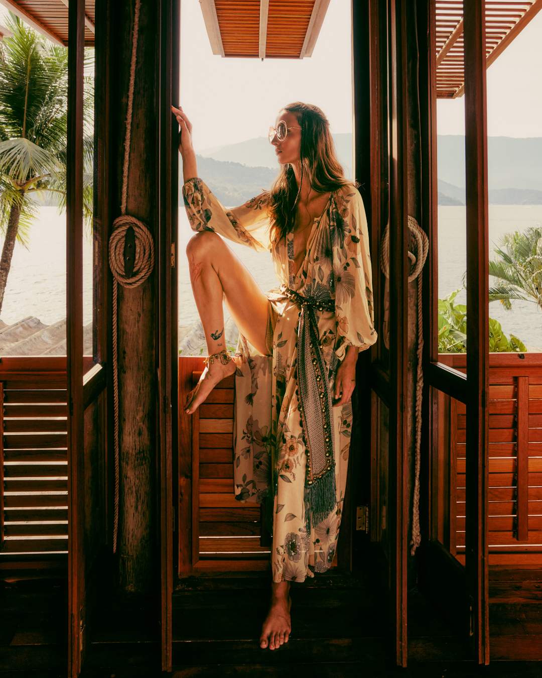Modelo em vestuário floral leve, posando em varanda com vista tropical. Tendências de alto verão.