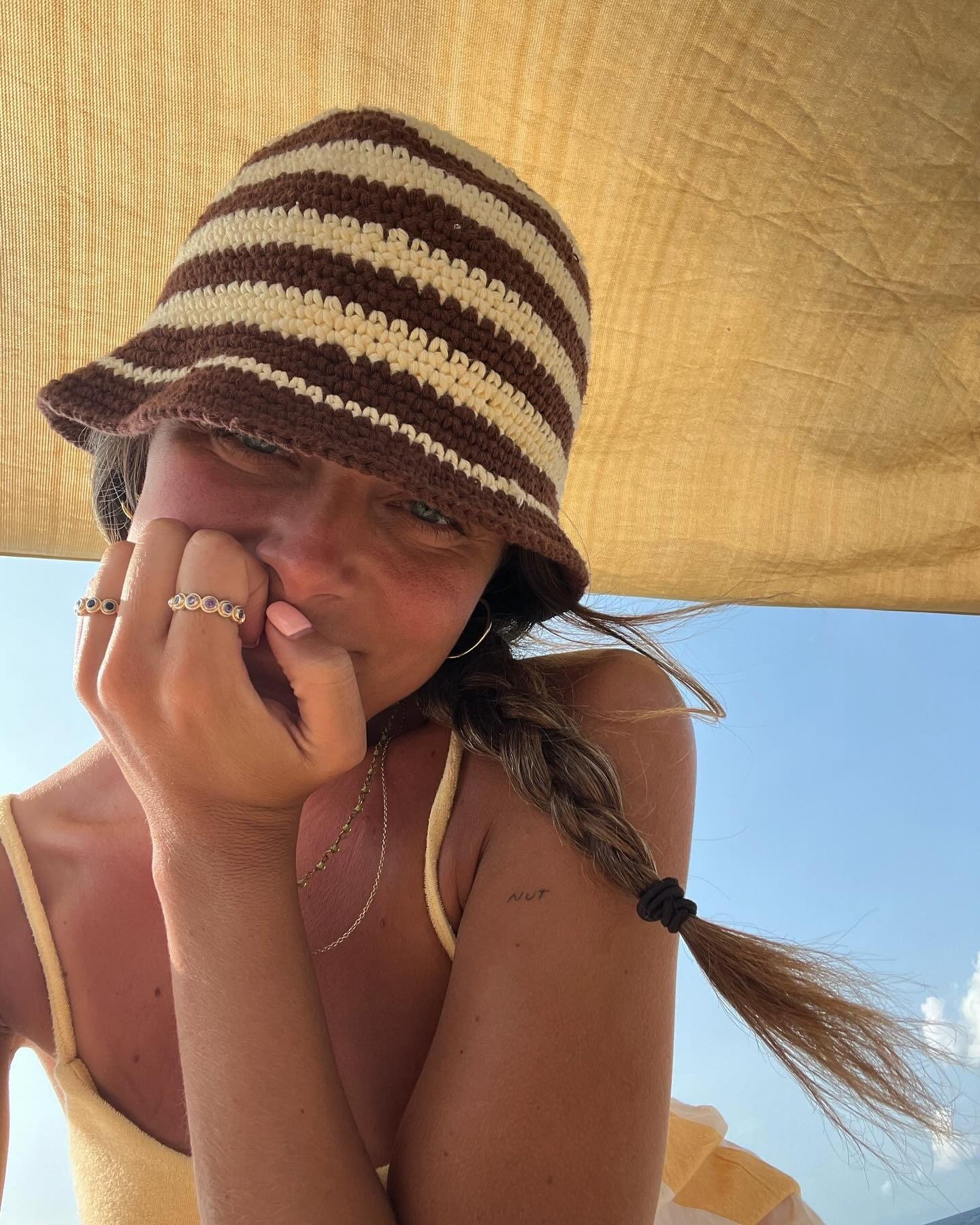 A imagem destaca uma jovem com um chapéu de crochê listrado em tons de marrom e bege, uma tendência de chapéu que combina estilo e proteção solar. Ela usa um vestido amarelo, com acessórios delicados como anéis e brincos, evidenciando um look casual e sof