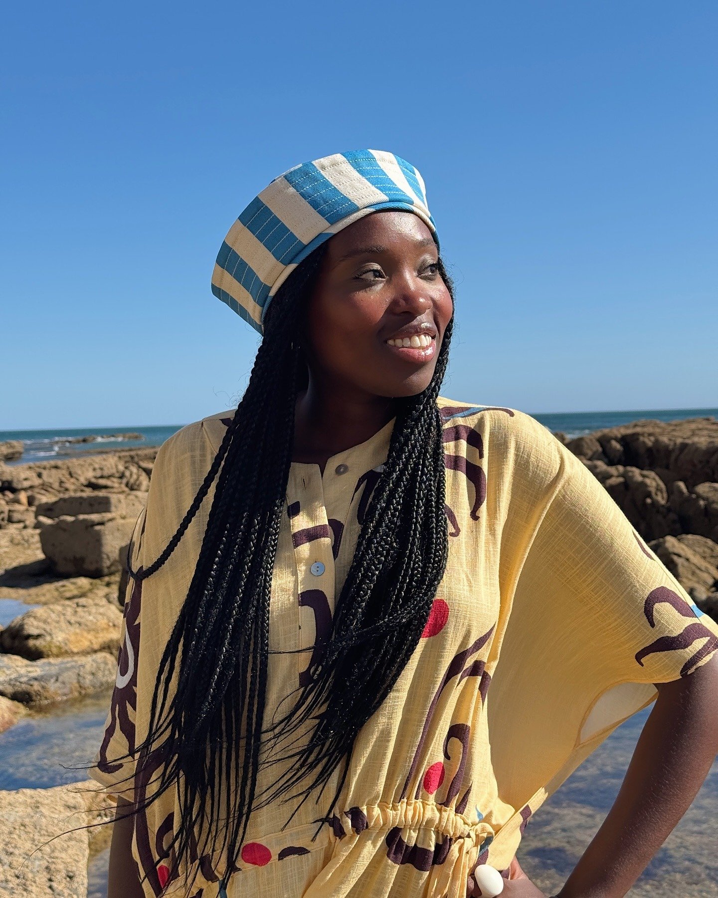 Com ares de verão, a imagem destaca uma mulher usando um chapéu listrado em azul e branco, que complementa seu vestido amarelo leve e estampado. A combinação de cores vibrantes e o cenário à beira-mar realçam as tendências de chapéu, que estão em alta na